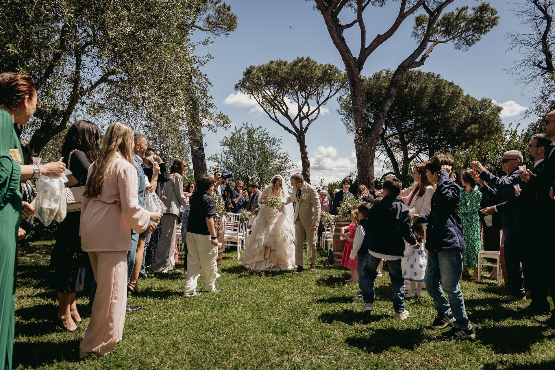 Gli sposi camminano su un sentiero erboso, mentre gli invitati lanciano coriandoli in una giornata di sole.