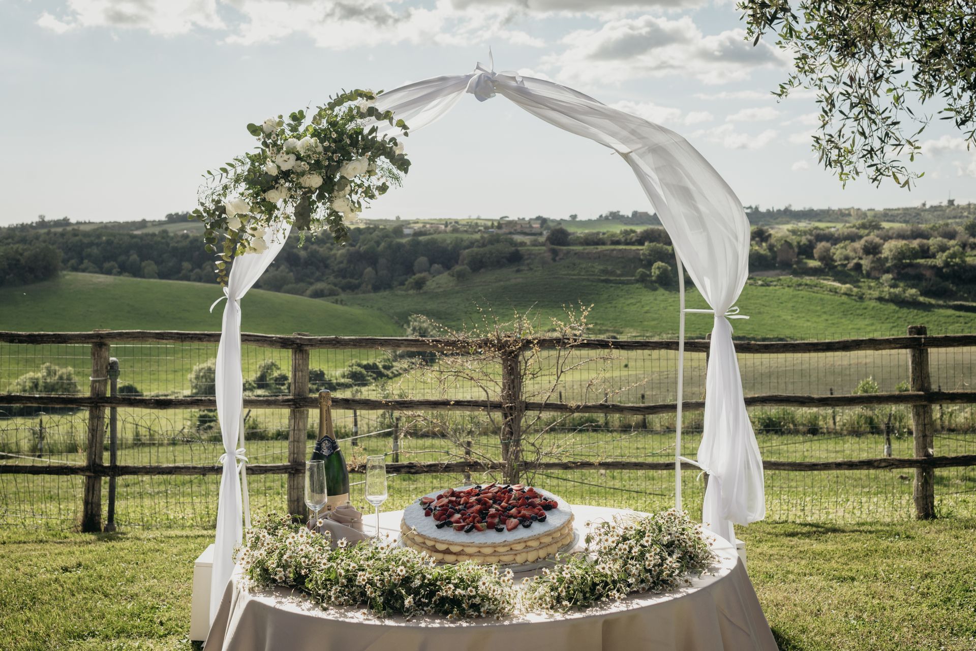 Tavolo per torte all'aperto con arco, fiori e dolci colline verdi sullo sfondo.