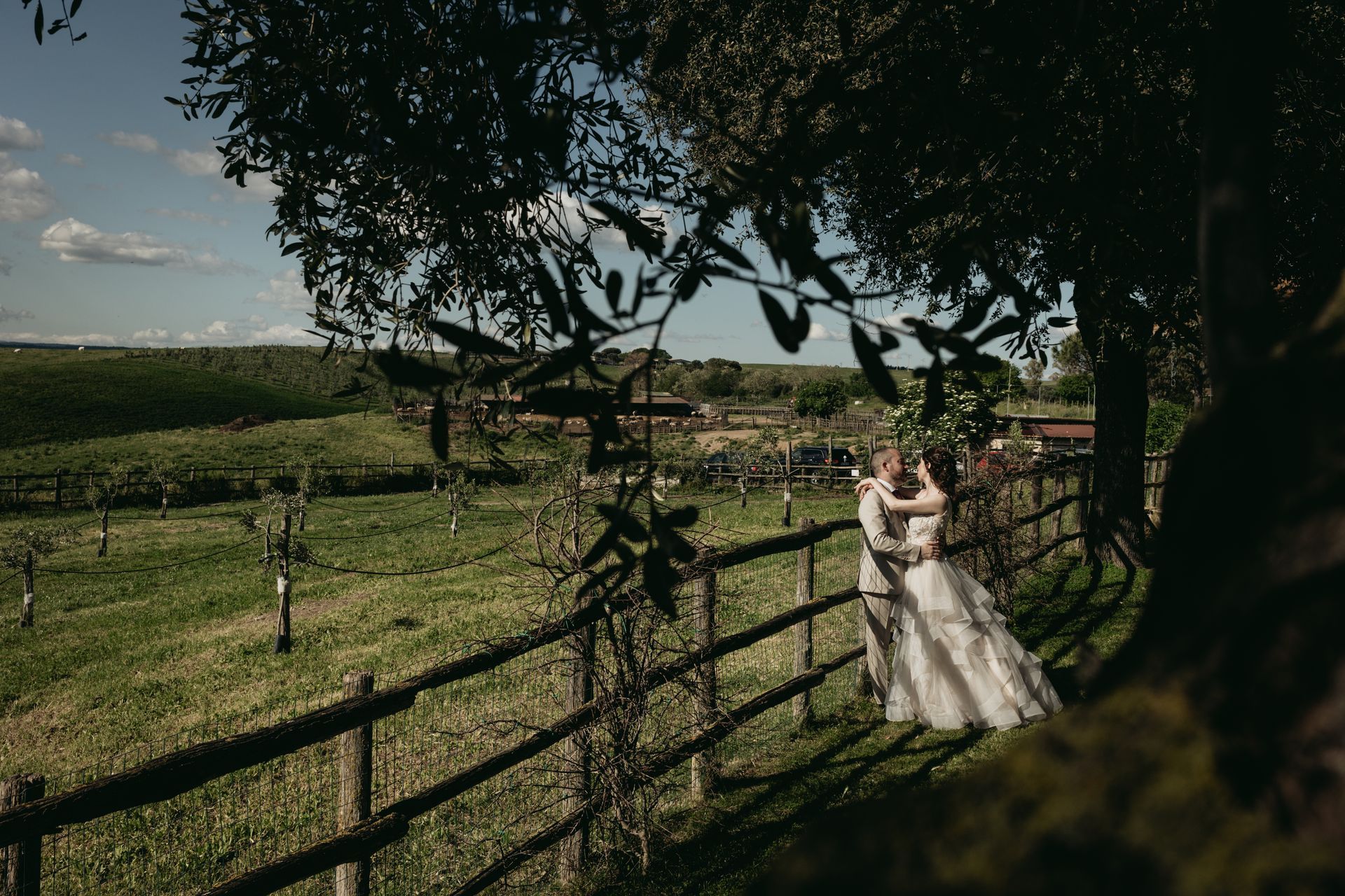 Coppia abbracciata vicino a una staccionata di legno, sullo sfondo una pittoresca campagna con campi verdi, giornata di sole.
