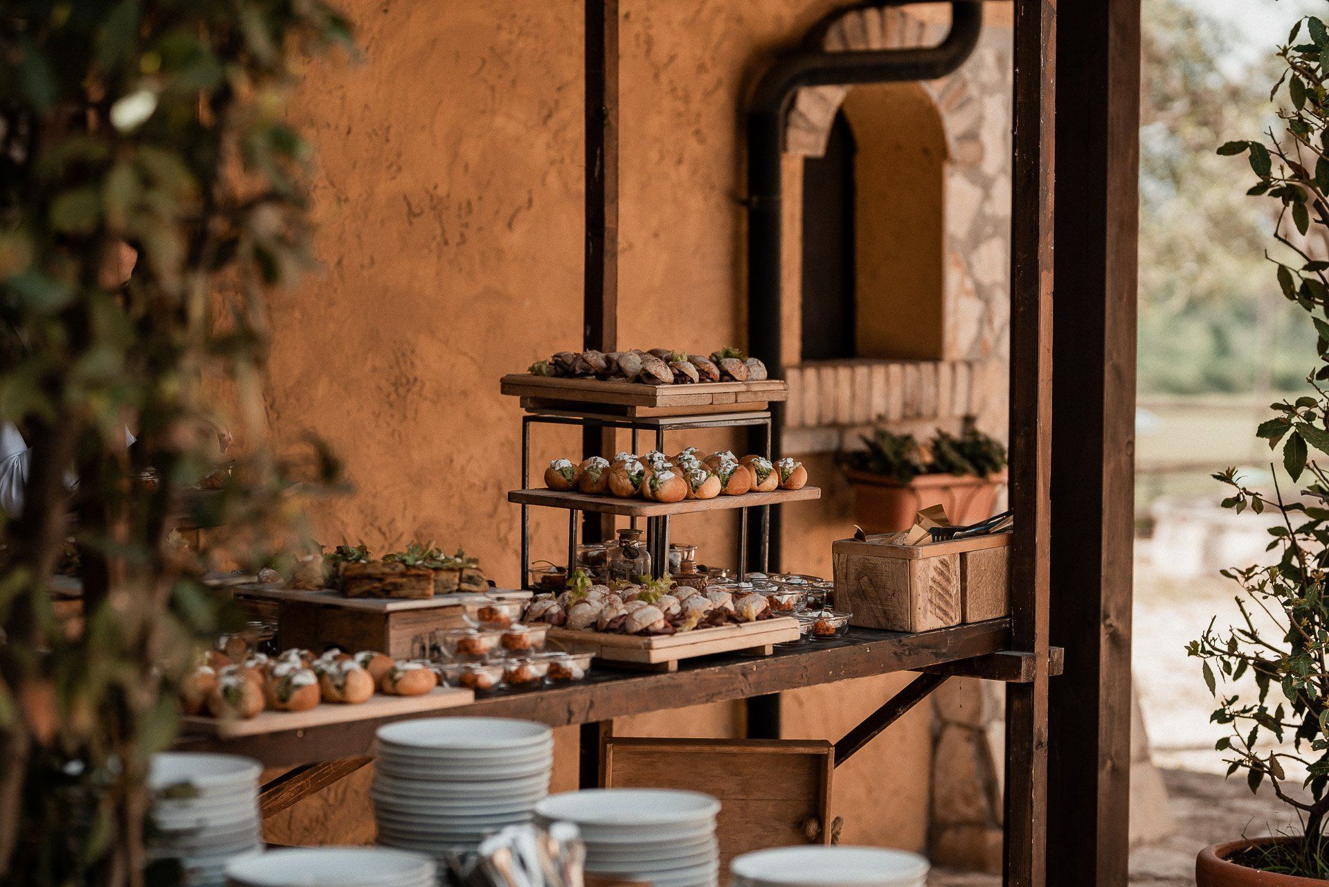 Tavolo da buffet con vassoi di antipasti disposti a più livelli e piatti bianchi impilati, all'aperto.