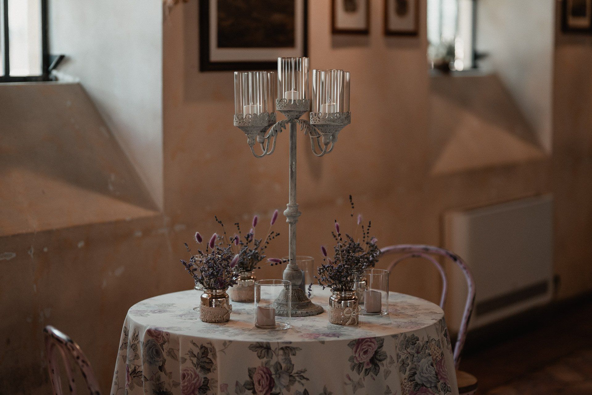 Apparecchiatura della tavola con candelabri e piccole composizioni floreali, in una stanza con una finestra e un termosifone.