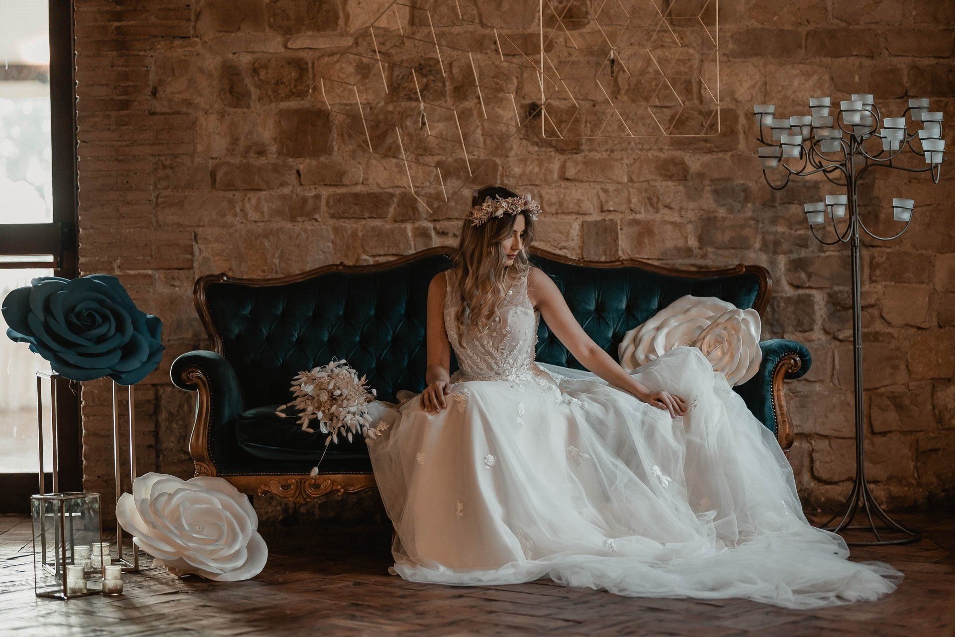 La sposa, in abito bianco, è seduta su un divano verde acqua, con in mano l'abito. Grandi fiori di carta e candele decorano la location.