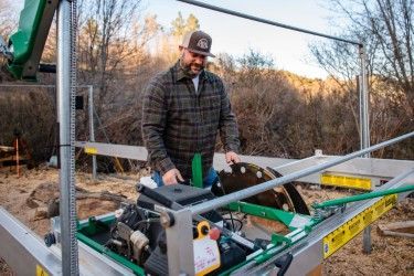 A person in a plaid shirt and cap operates a green portable sawmill outdoors among trees.