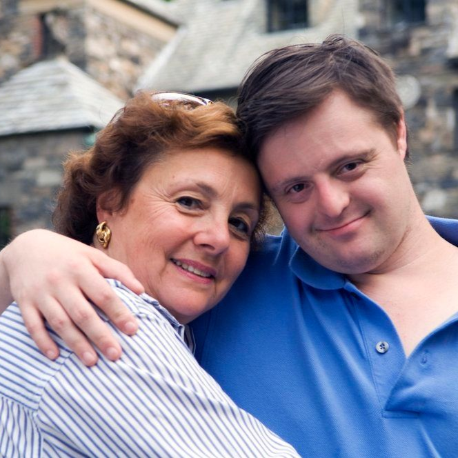 Woman with arm around person with Down syndrome, smiling, near a stone building.