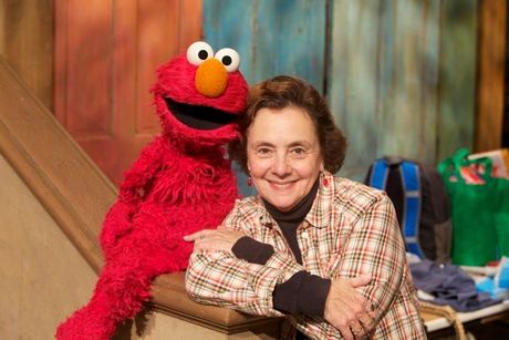 Elmo the Muppet with a woman, smiling, in a plaid shirt on the Sesame Street set.
