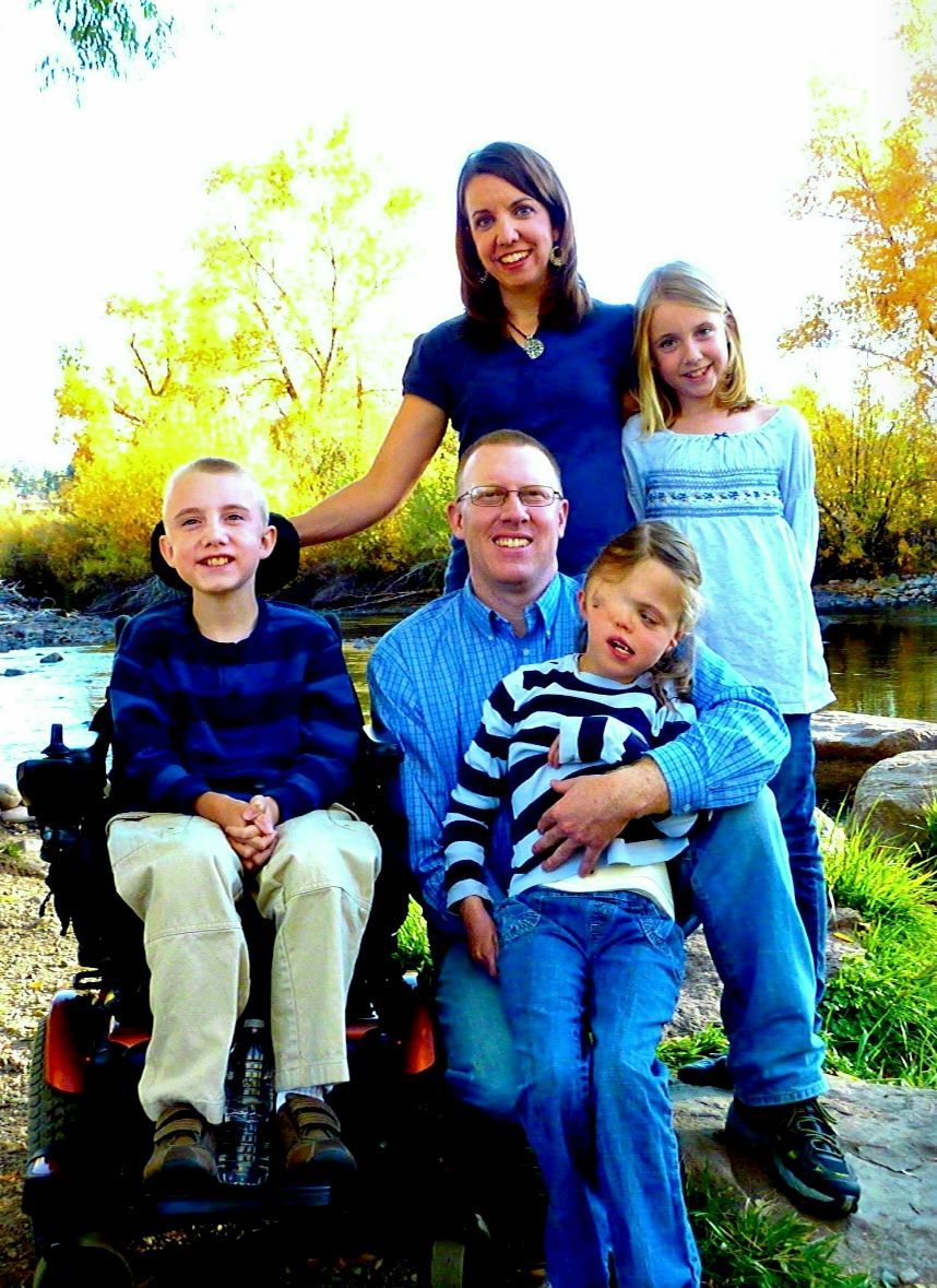 Family posing outdoors by a river. Mother and daughter standing, father with two children sitting in his lap and in a wheelchair.