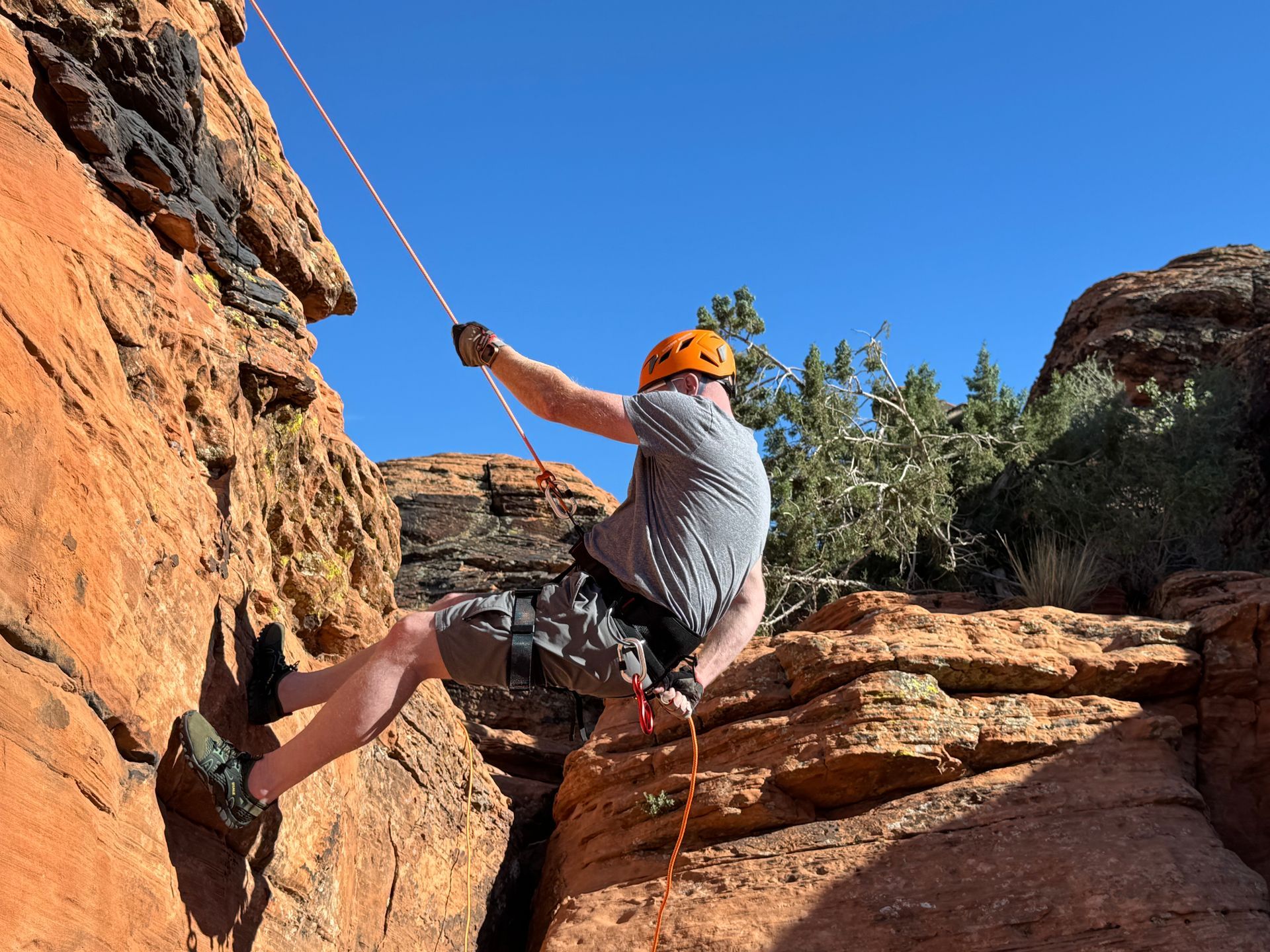 Man rappelling down a red rock cliff, wearing a helmet and harness, sunny day.