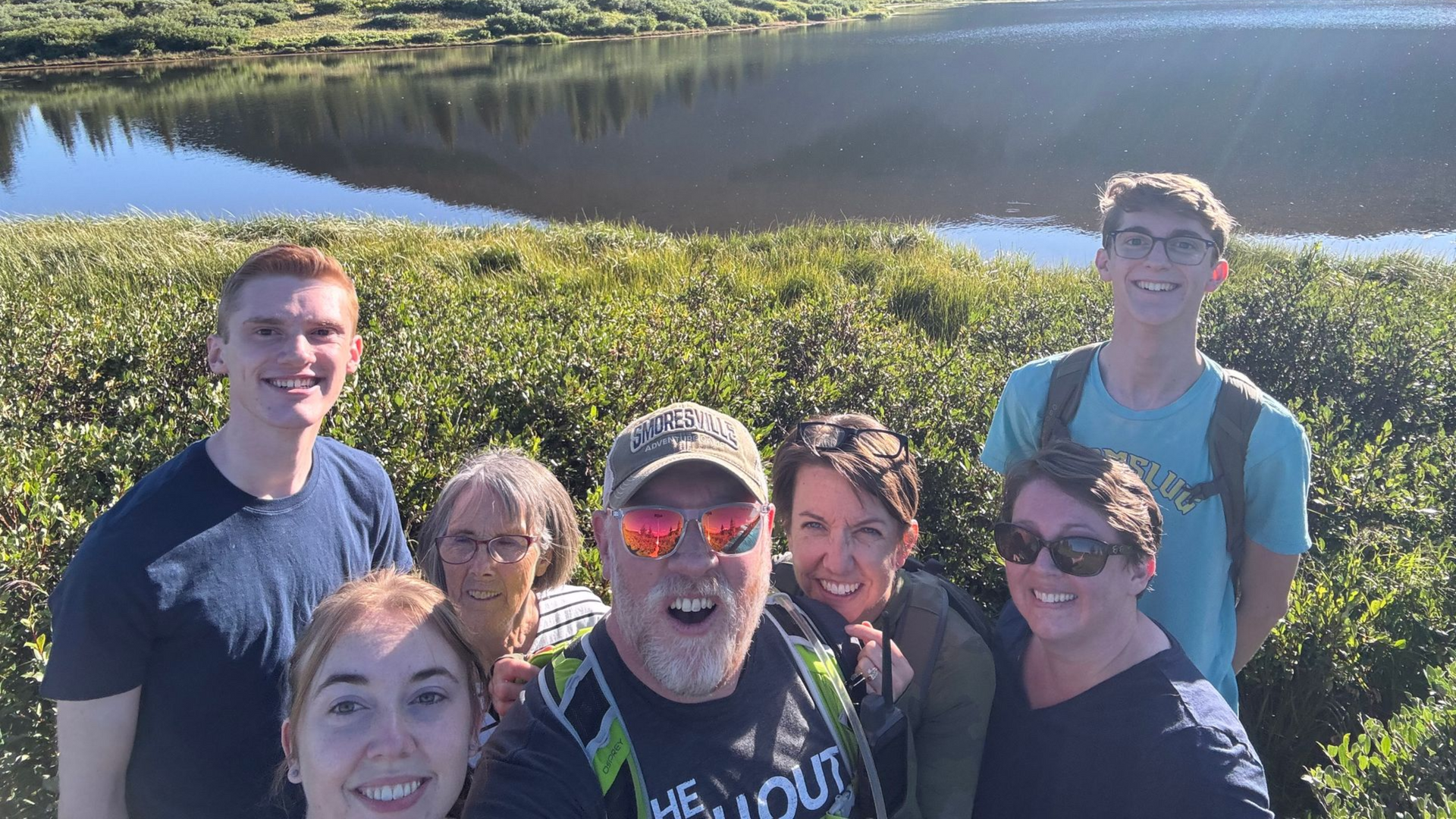 Group selfie near a reflective lake. Seven people smile, framed by green bushes; sunny, outdoor setting.