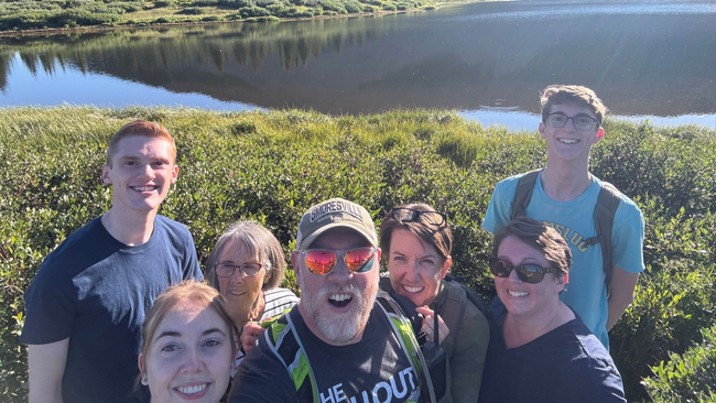 Group selfie near a reflective lake. Seven people smile, framed by green bushes; sunny, outdoor setting.