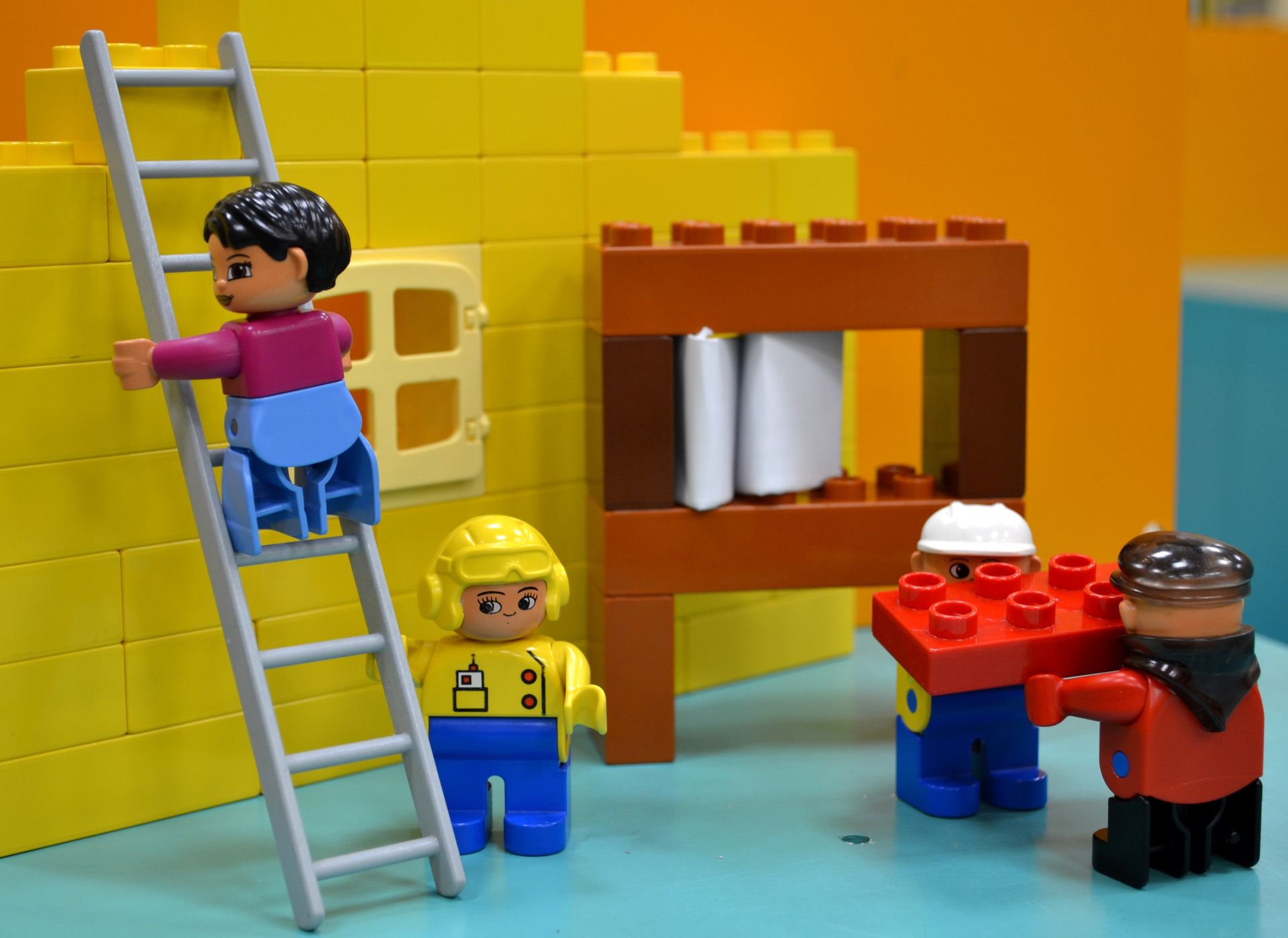 Lego figures building a structure: one climbing ladder, others holding a red brick, and one observing near yellow brick wall.