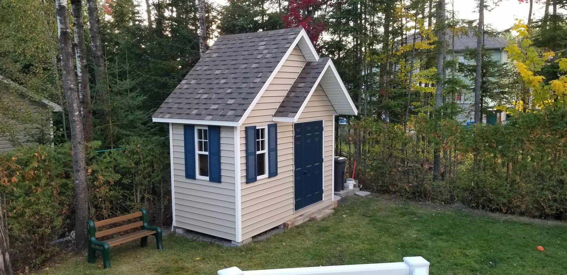 Une petite cabane en bois avec des volets bleus, une porte sombre et un toit de bardeaux bruns, située dans une cour.