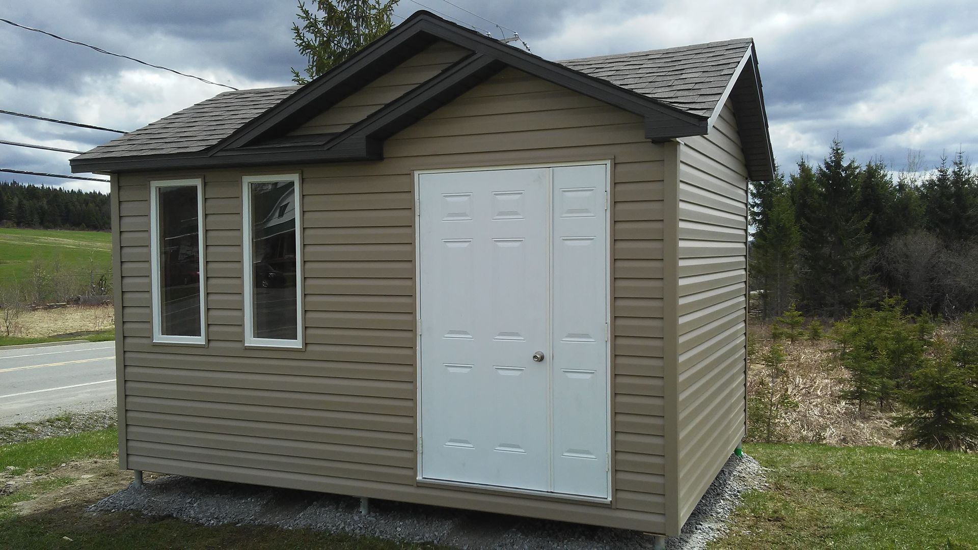 Abri de jardin marron avec porte blanche et deux fenêtres, situé dans une cour herbeuse.