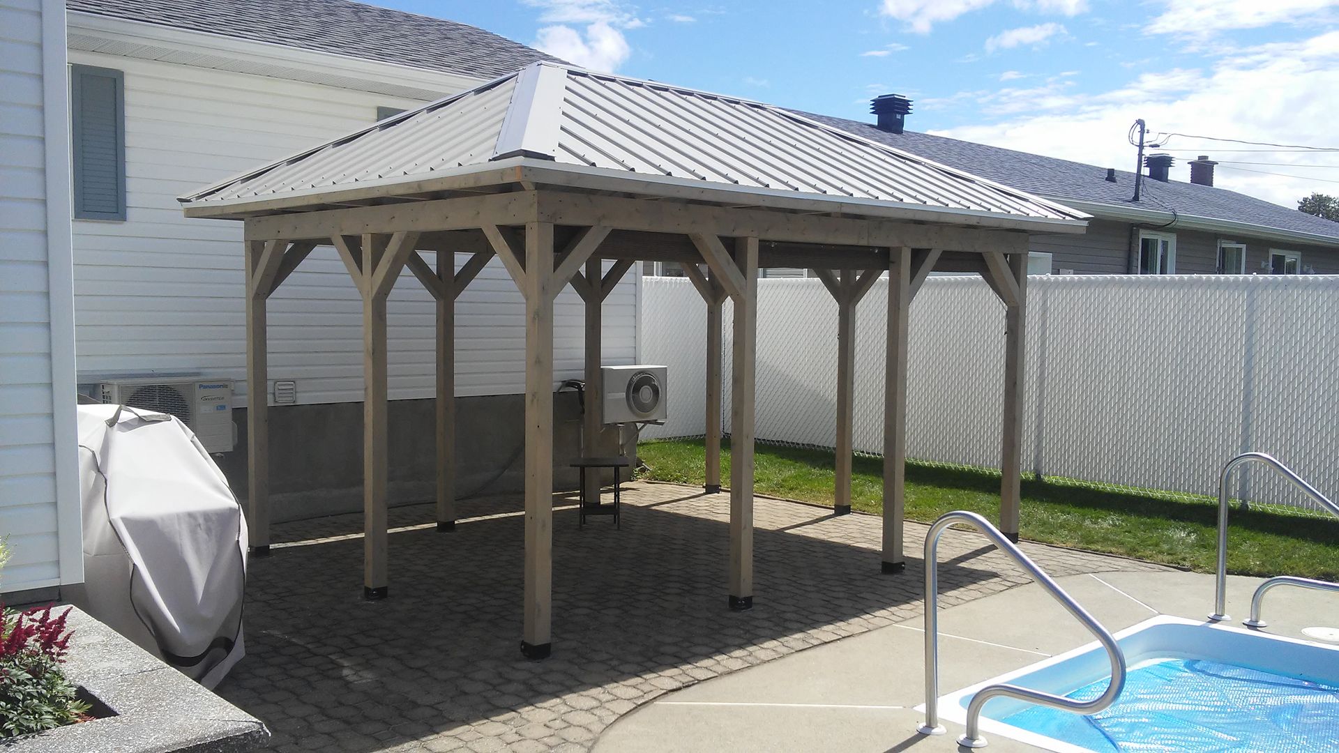 Gazebo beige avec toit en métal au-dessus d'une terrasse en briques à côté d'une piscine, dans le jardin.