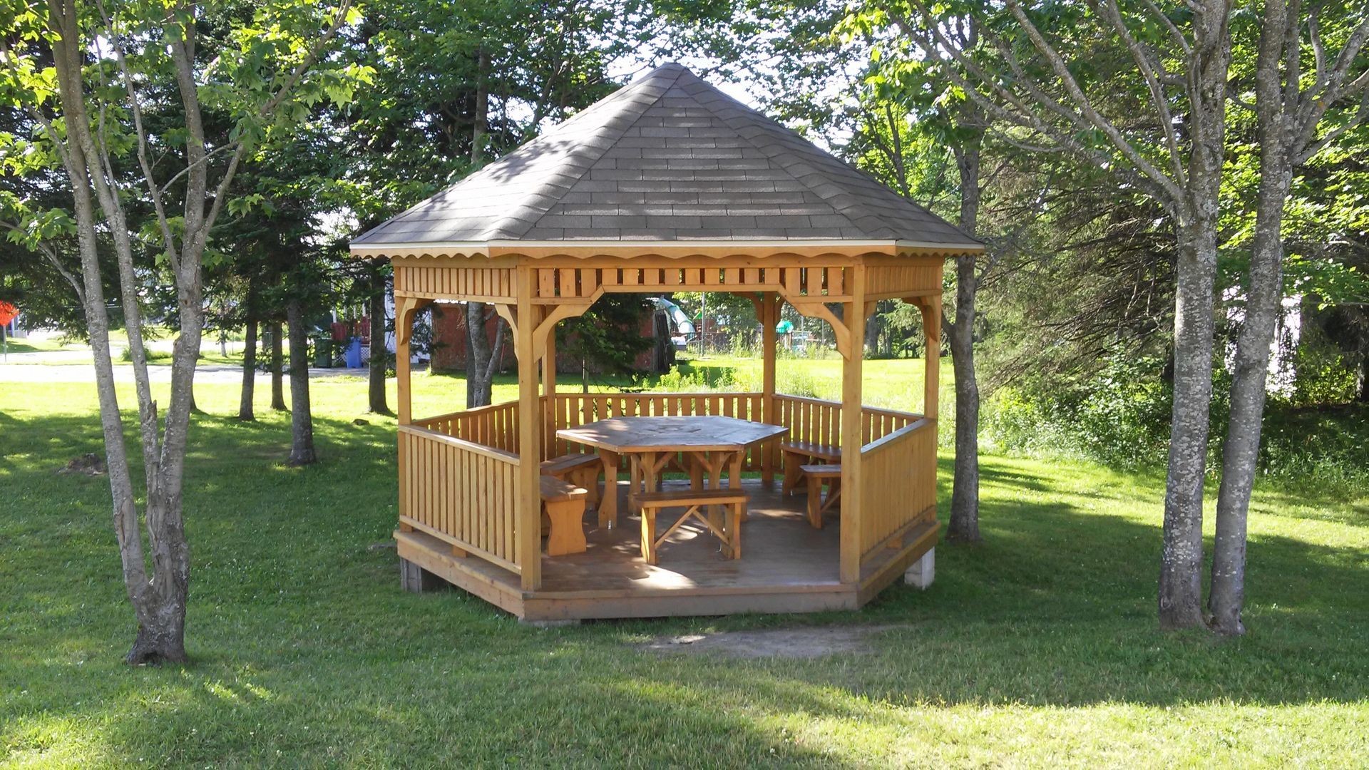 Un kiosque en bois avec une table et des bancs se trouve sur une pelouse entourée d'arbres.