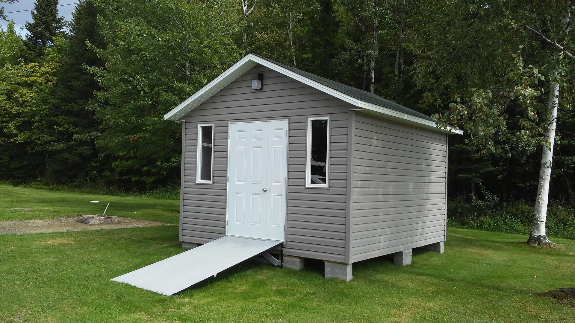 Abri de jardin gris avec porte blanche, rampe et fenêtres, sur une pelouse arborée.