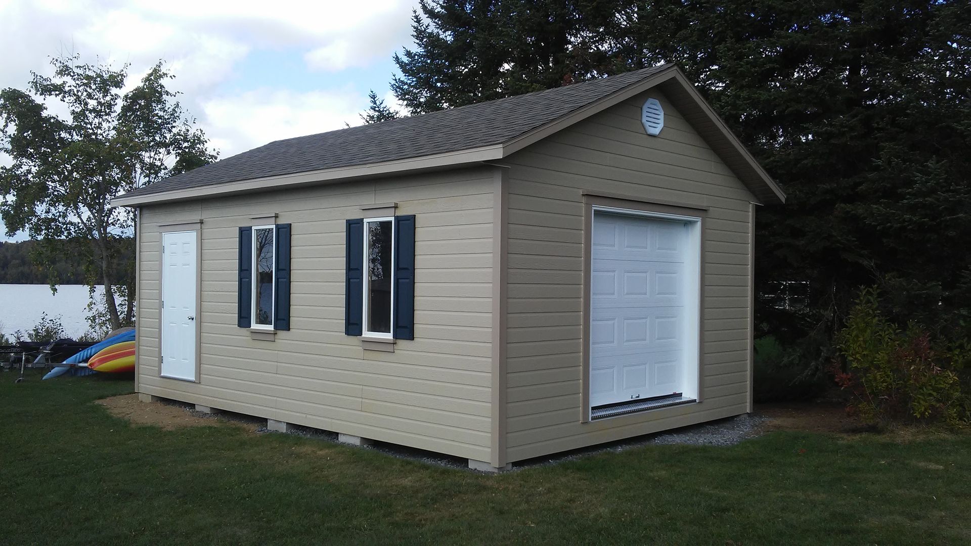 Abri de jardin beige avec porte de garage, porte latérale et deux fenêtres à volets bleus, installé sur l'herbe.