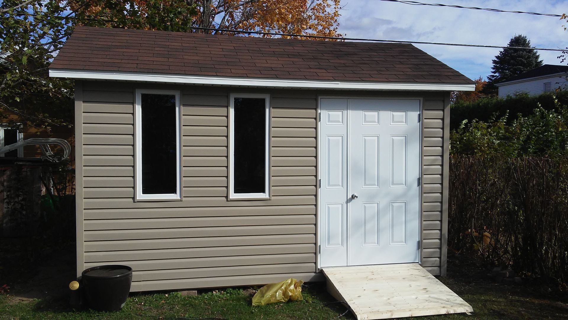 Abri beige avec toit marron, deux fenêtres, porte blanche avec rampe.