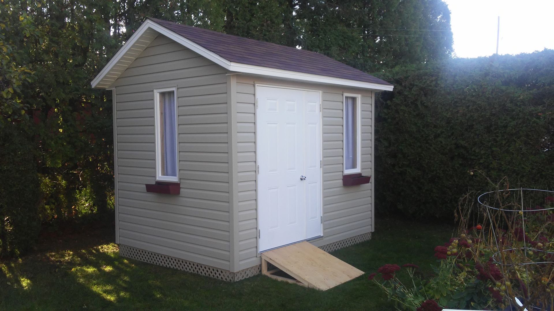 Abri beige avec porte et fenêtres blanches, toit marron, rampe en bois sur herbe verte.