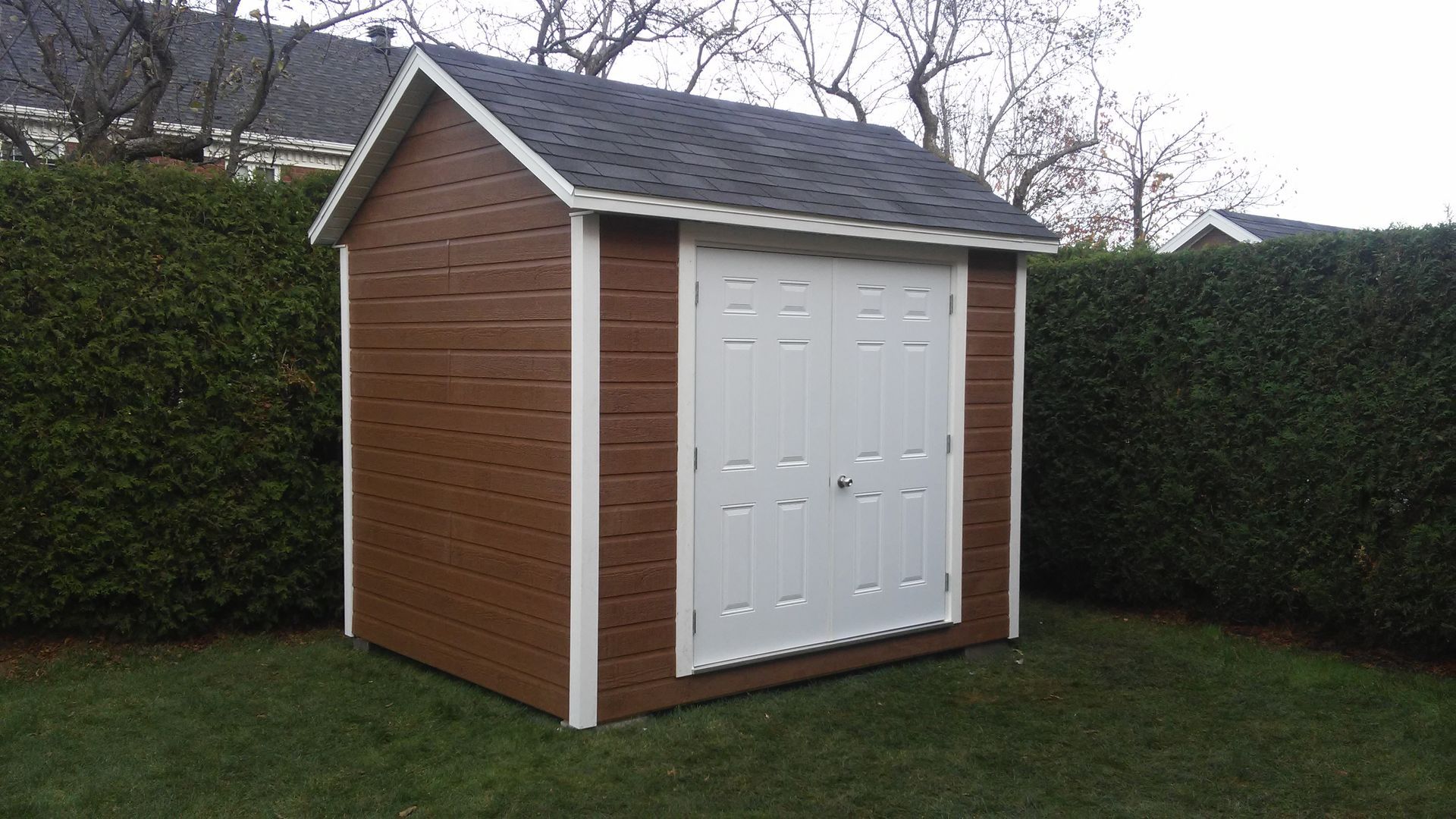 Abri de jardin marron avec portes et encadrements blancs, toit noir, sur une pelouse verte.