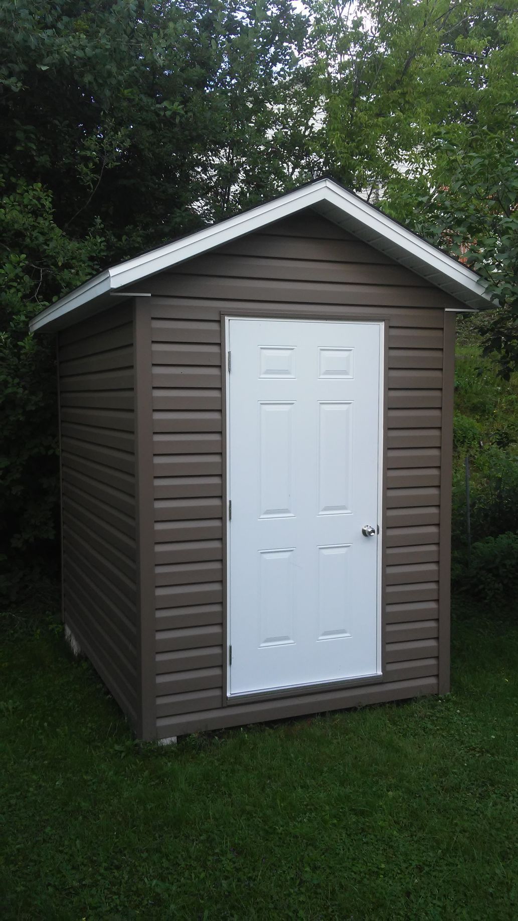 Cabane marron à porte blanche, située dans une pelouse verte avec des arbres en arrière-plan.