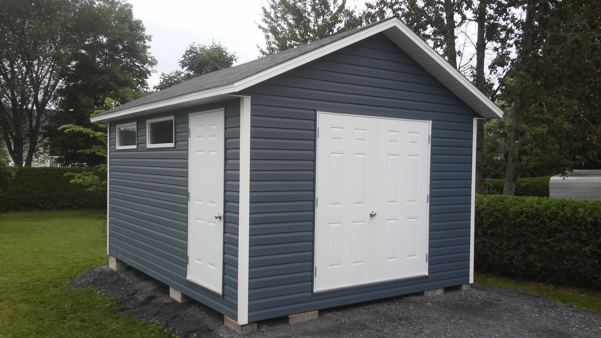 Abri de jardin bleu foncé avec portes et encadrements blancs, sur un sol en gravier, situé dans une cour.