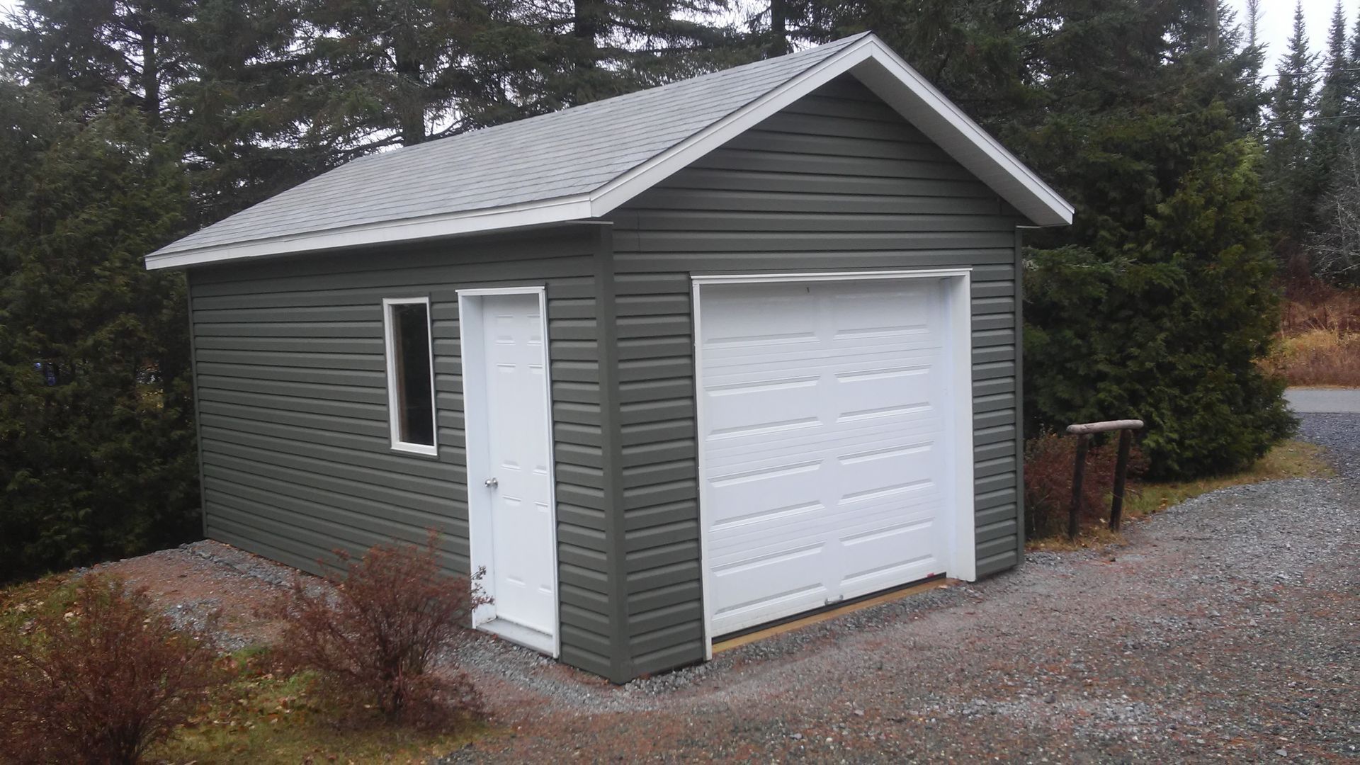 Petit garage vert avec porte blanche. Allée en gravier.