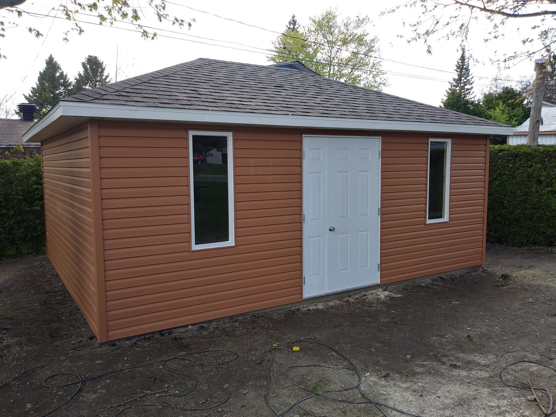 Abri de jardin marron avec fenêtres à cadre blanc et portes doubles, toit en bardeaux gris, situé dans une cour.