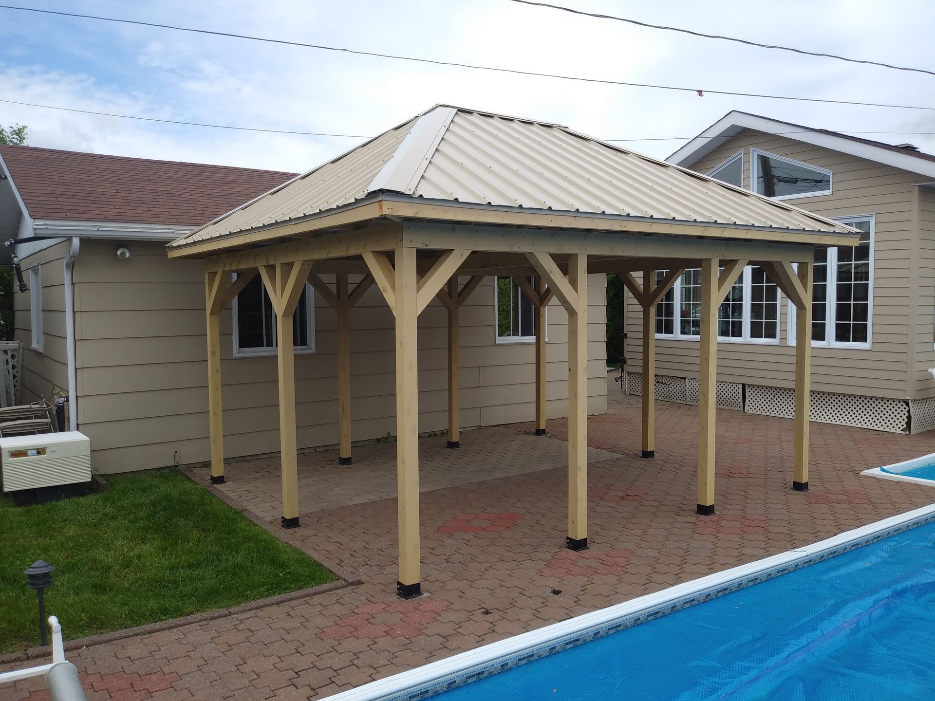 Gazebo en bois au toit beige, construit au-dessus d'une terrasse en briques, à côté d'une piscine et d'une maison aux murs beiges.