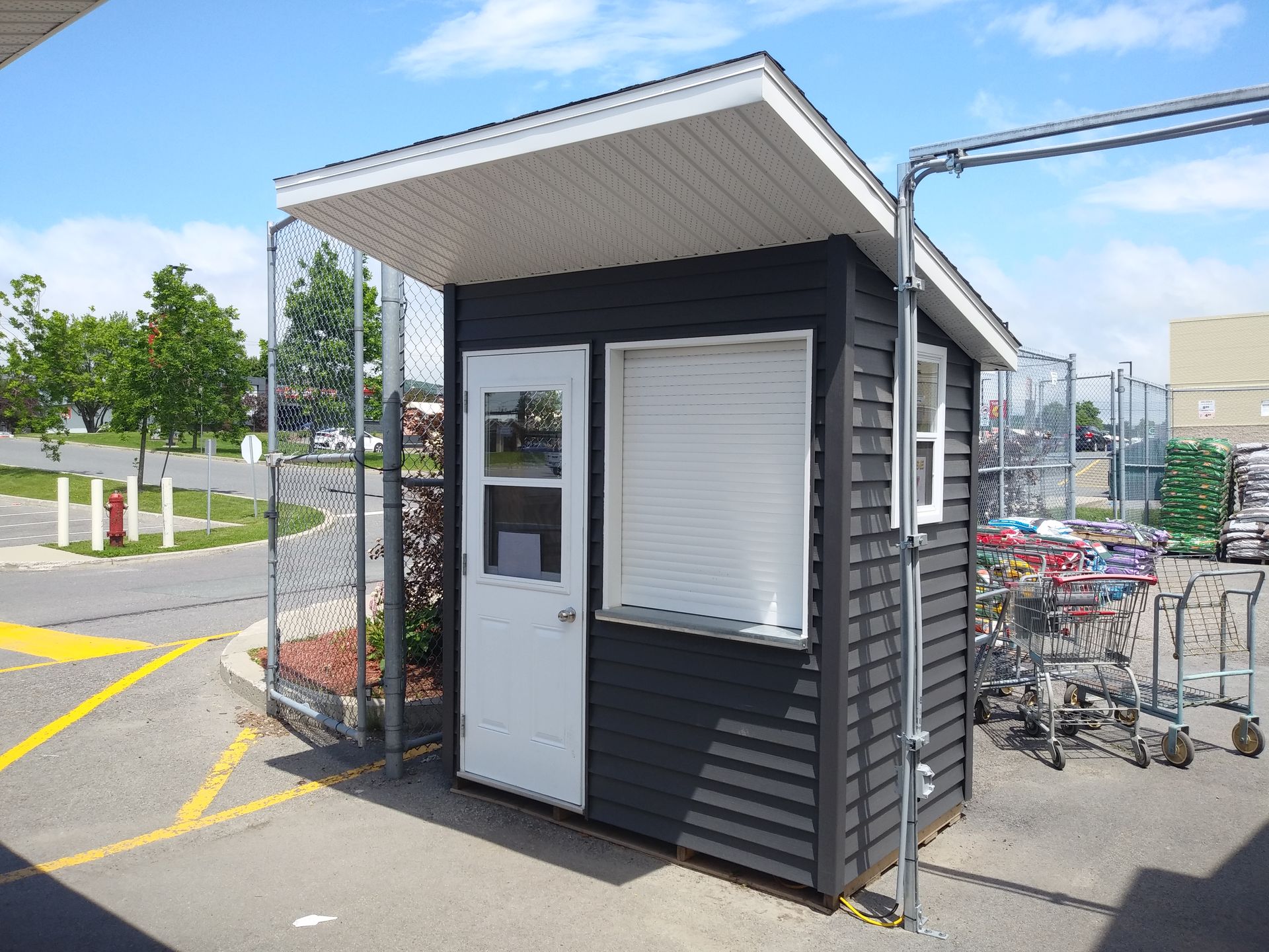 Cabine gris foncé dans un enclos à chariots de supermarché extérieur, avec porte et fenêtre blanches, sous un toit blanc.