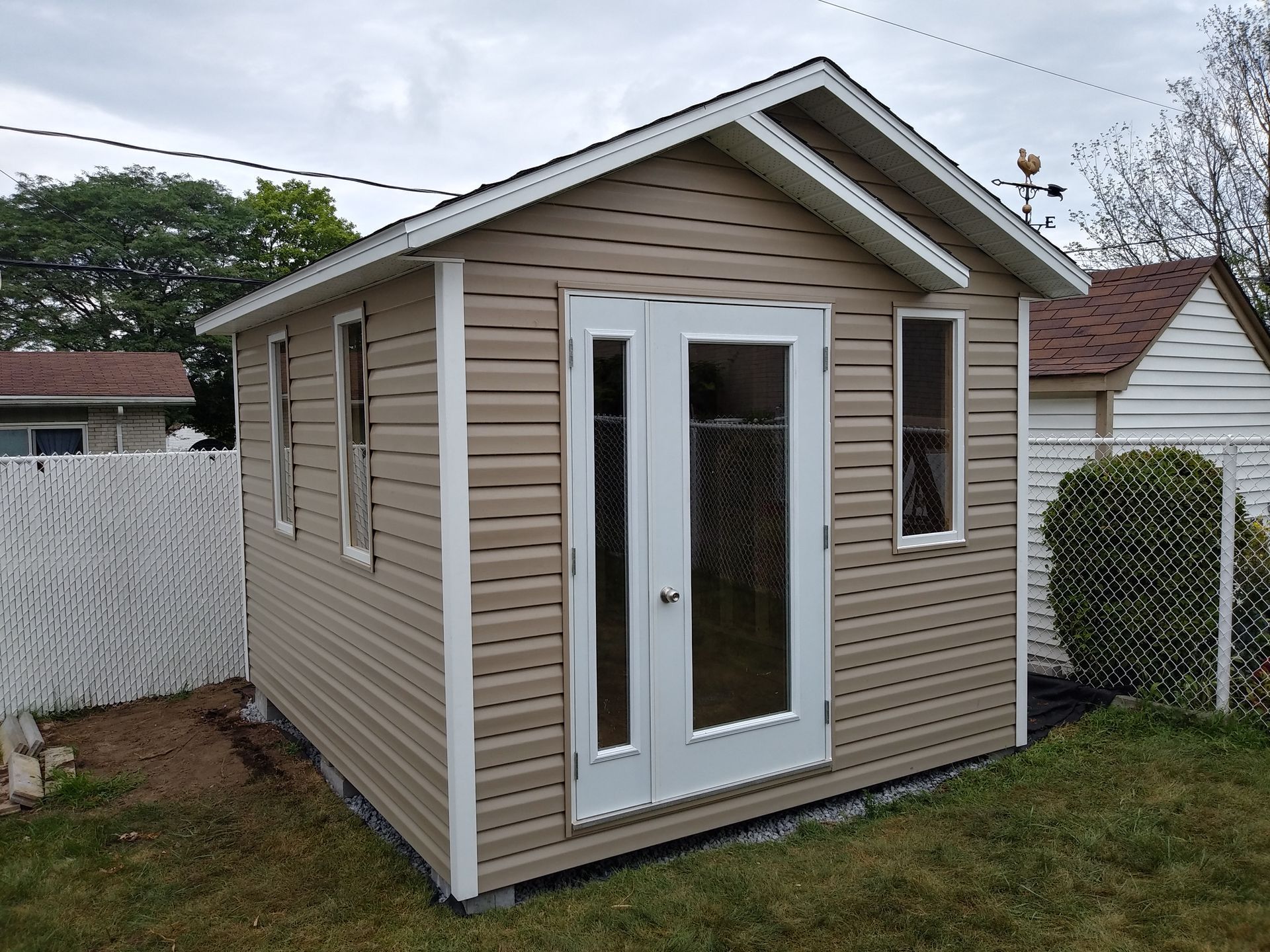 Abri de jardin beige avec des finitions blanches, des portes doubles et deux fenêtres dans une cour.