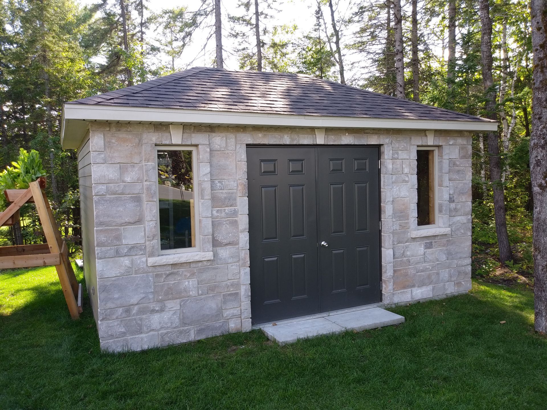Abri en parpaings avec double porte, deux fenêtres et toit en bardeaux gris, entouré d'herbe verte et d'arbres.