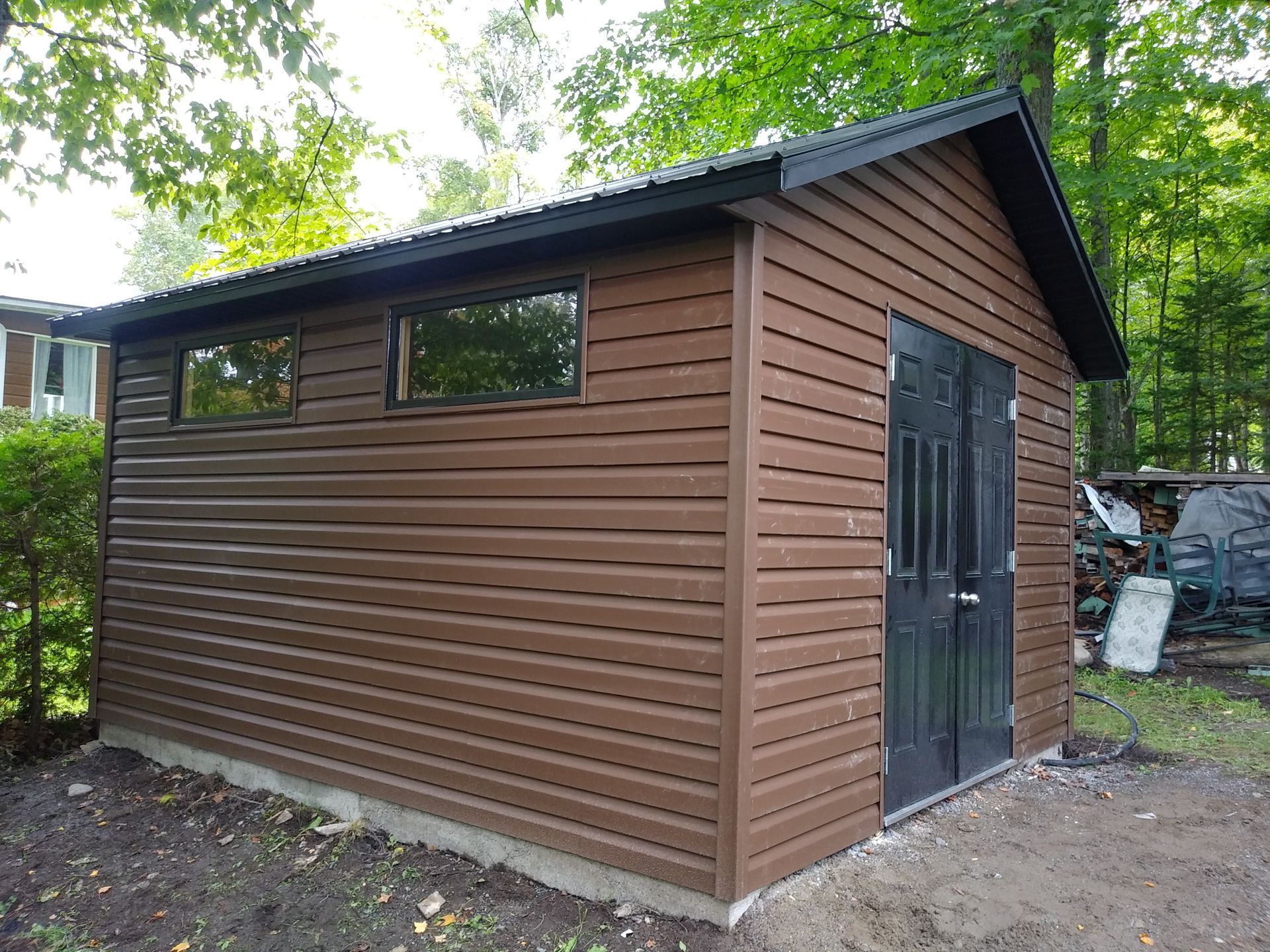 Abri marron avec boiseries et portes noires, deux fenêtres, toit en tôle ondulée, arbres en arrière-plan.