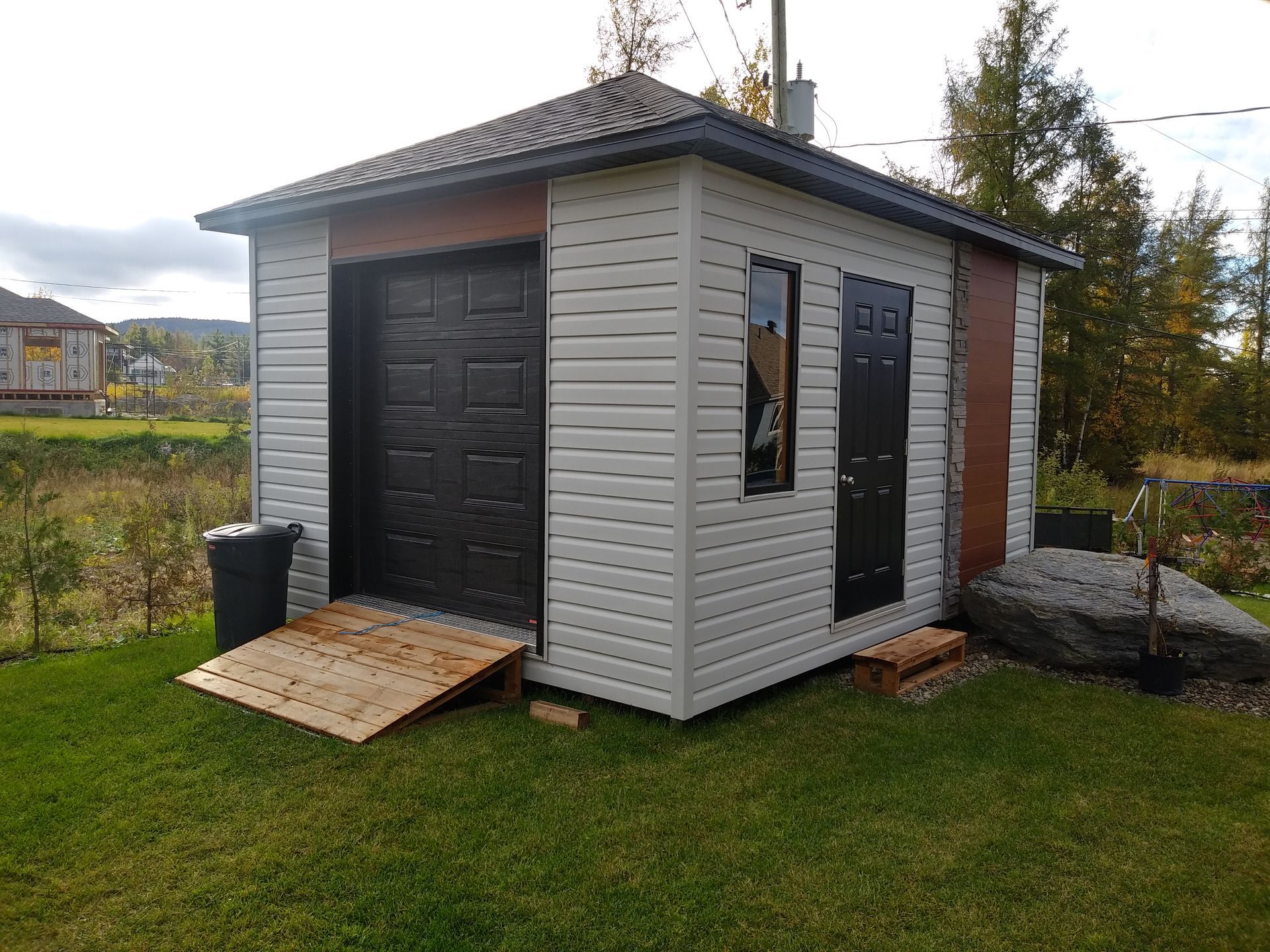 Petit abri avec porte noire, rampe et porte latérale, sur pelouse verte.