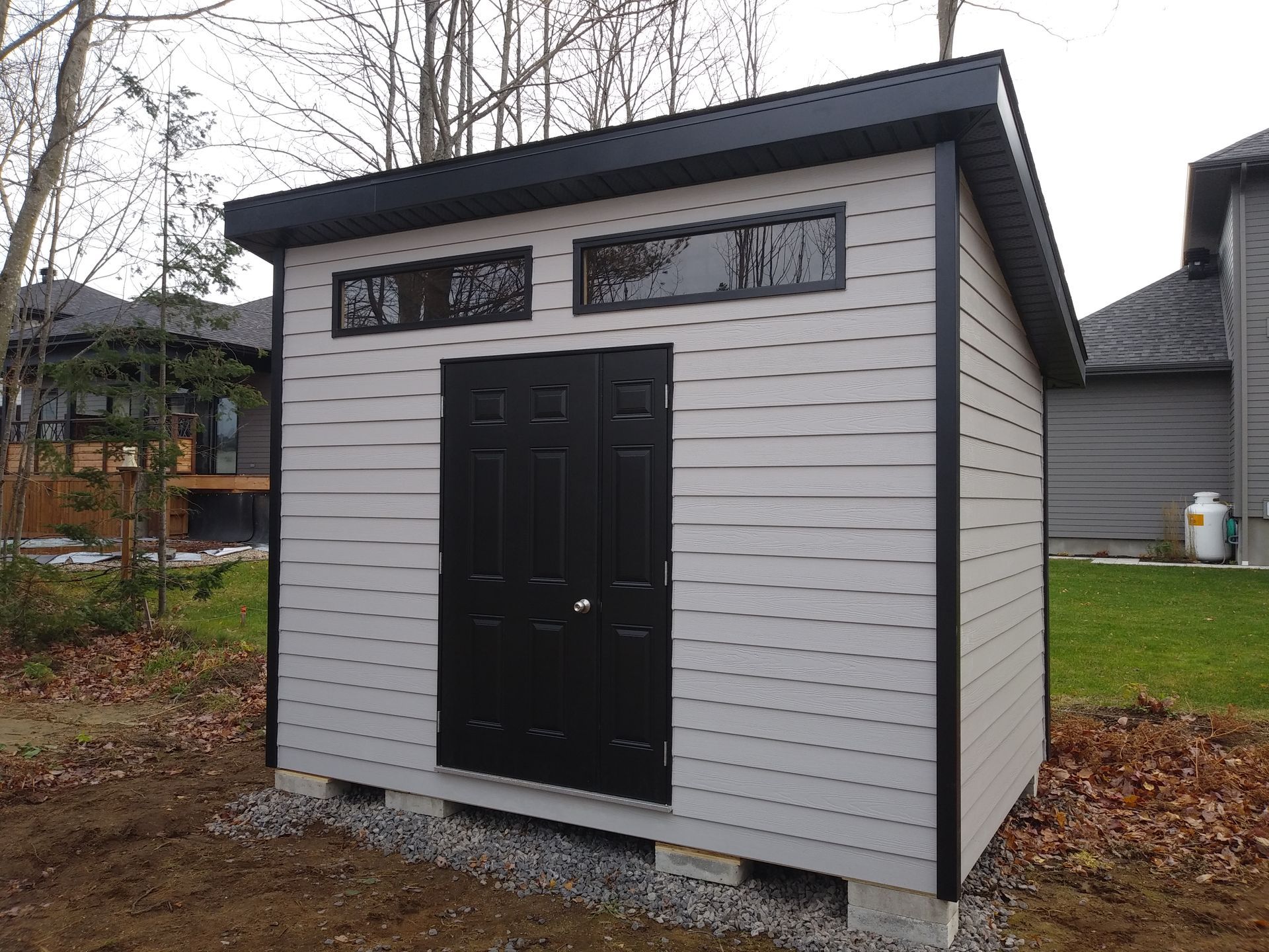 Abri de jardin gris avec porte et encadrement noirs, trois petites fenêtres, sur un sol en gravier.