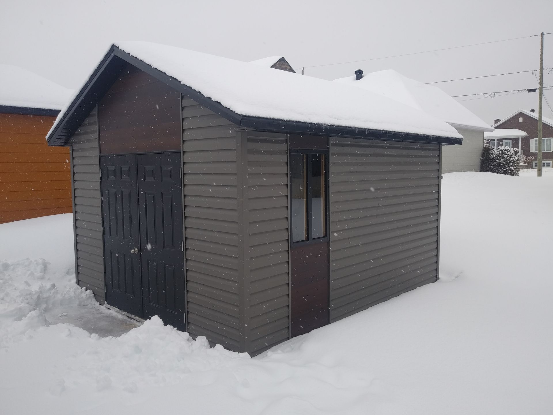 Petit hangar recouvert de neige, avec une porte et un bardage gris foncé, dans un environnement enneigé.