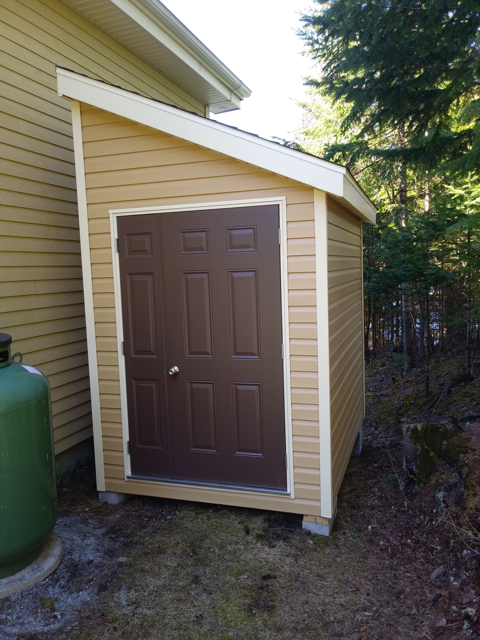 Un abri beige avec une porte marron, à côté d'une maison jaune et d'une bonbonne de propane verte, dans un cadre extérieur.