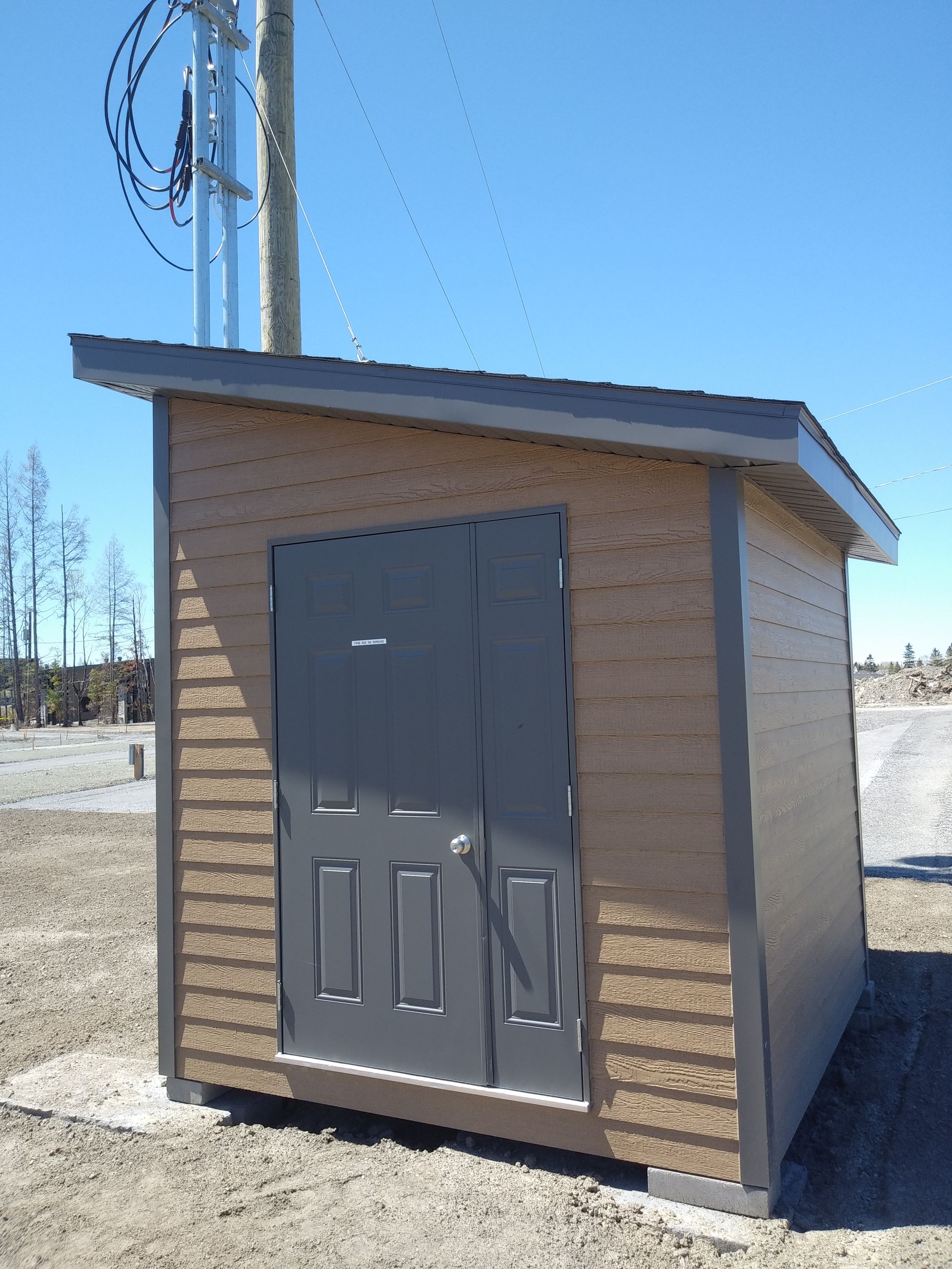 Abri de jardin beige et gris avec toit et porte en pente, situé près des lignes électriques.