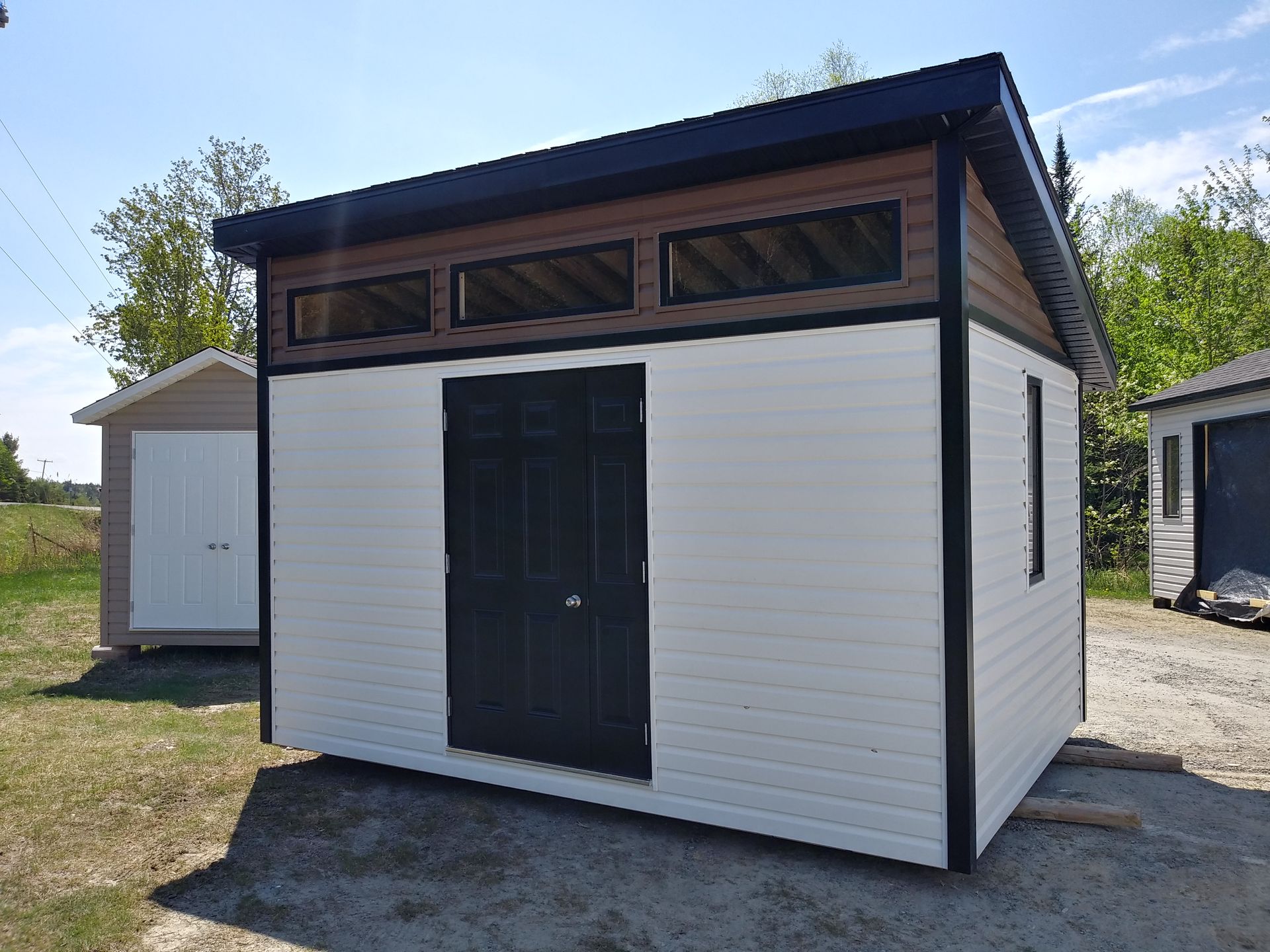 Abri de jardin blanc et marron avec des boiseries et une porte noires, trois petites fenêtres au-dessus de la porte.