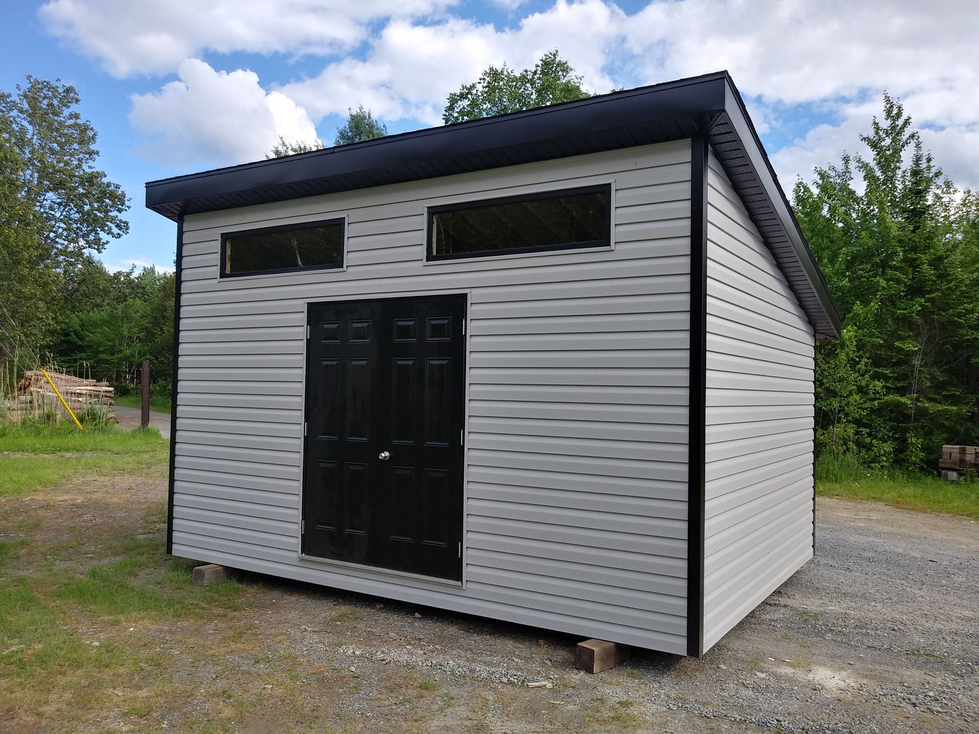 Abri de jardin gris et noir avec un toit en pente, deux fenêtres et des doubles portes noires, situé dans un espace herbeux.