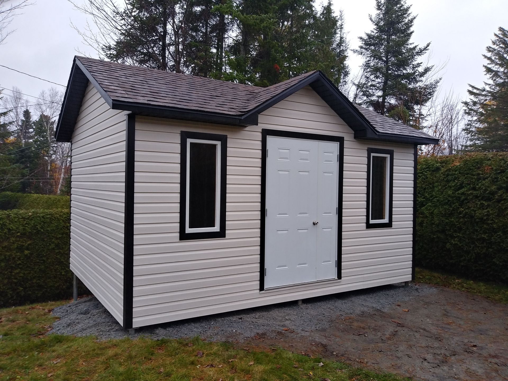 Abri de jardin beige avec des finitions noires et un toit marron, deux fenêtres et une porte blanche, posé sur du gravier.