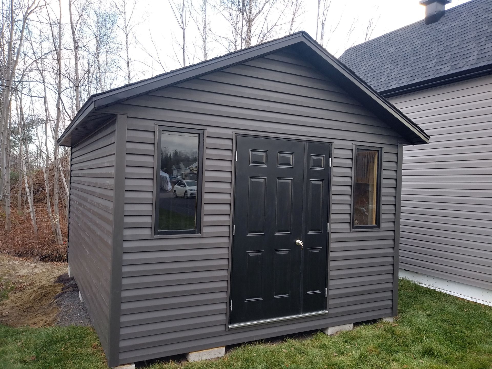 Un cabanon gris aux portes et encadrements de fenêtres noirs, situé sur une pelouse verte avec une maison en arrière-plan.