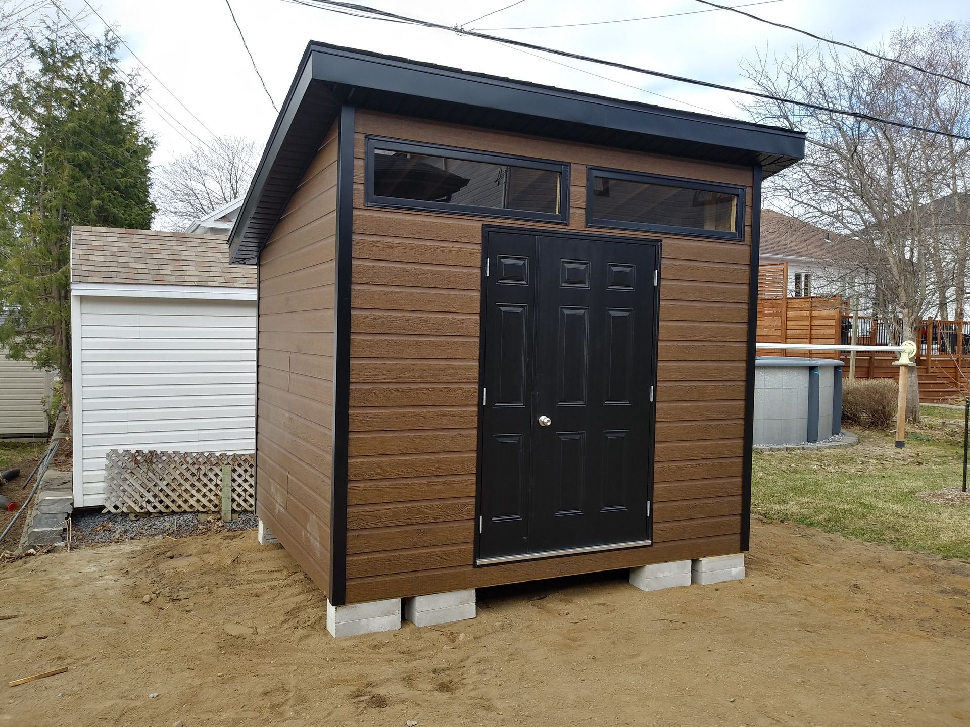 Abri de jardin marron avec porte, fenêtres et encadrements noirs, sur blocs de béton, à côté d'un abri de jardin blanc.