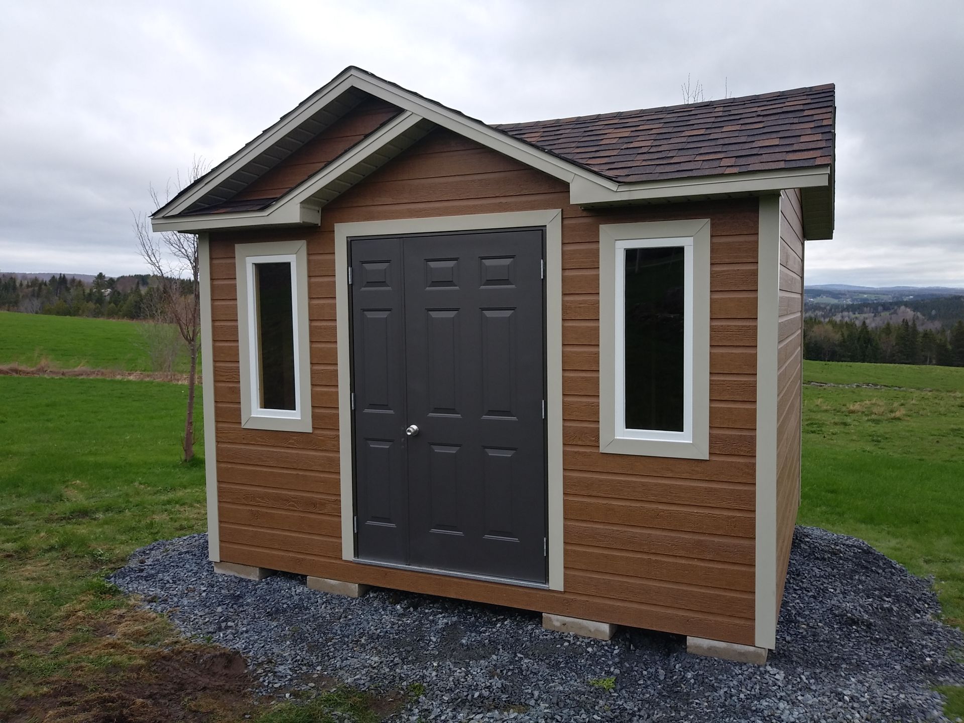 Un hangar marron avec une porte et des fenêtres sombres, sur un sol en gravier, situé dans un champ herbeux.