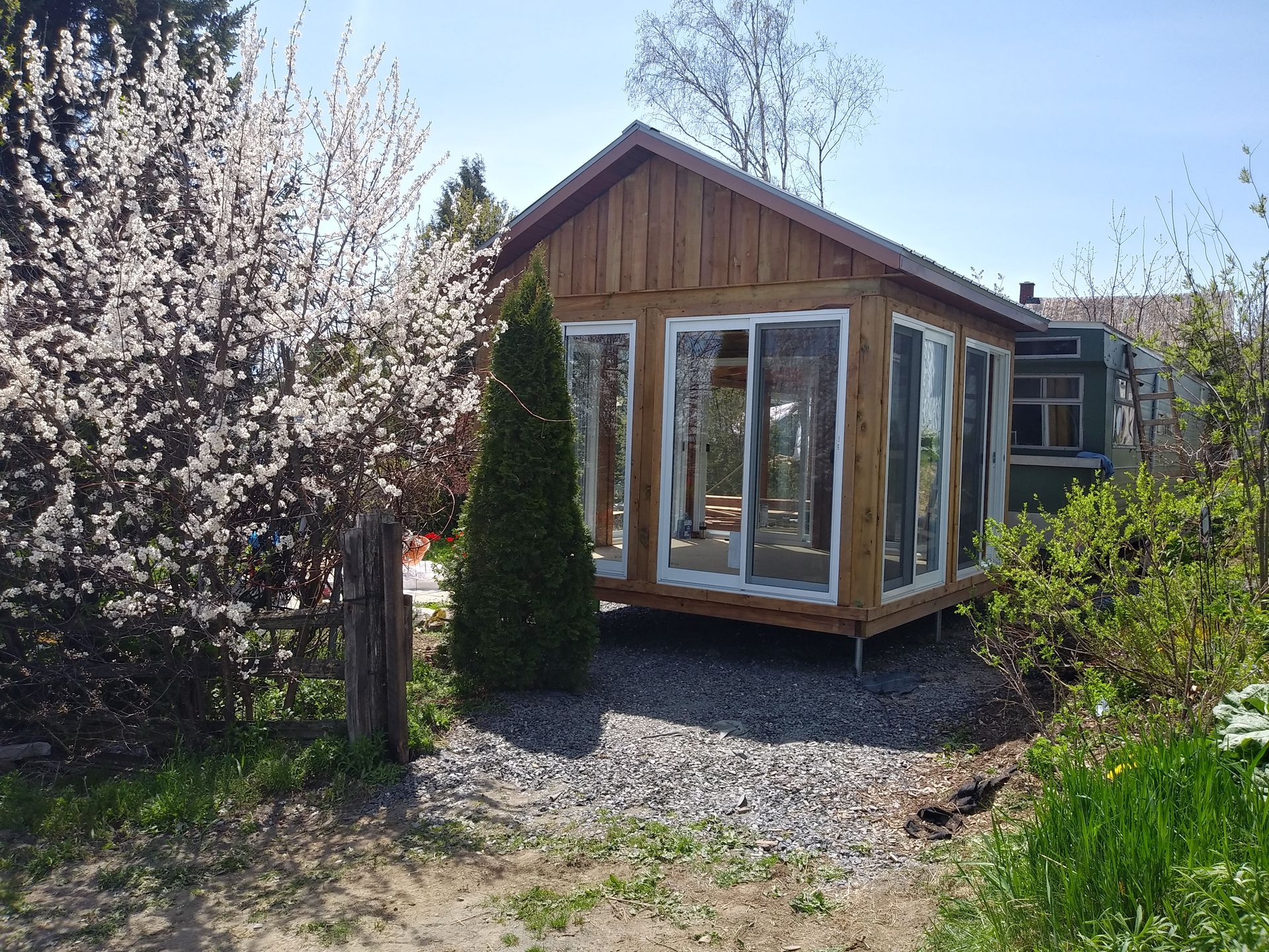 Véranda en bois avec de grandes fenêtres, à côté d'un arbre en fleurs et de buissons verdoyants.