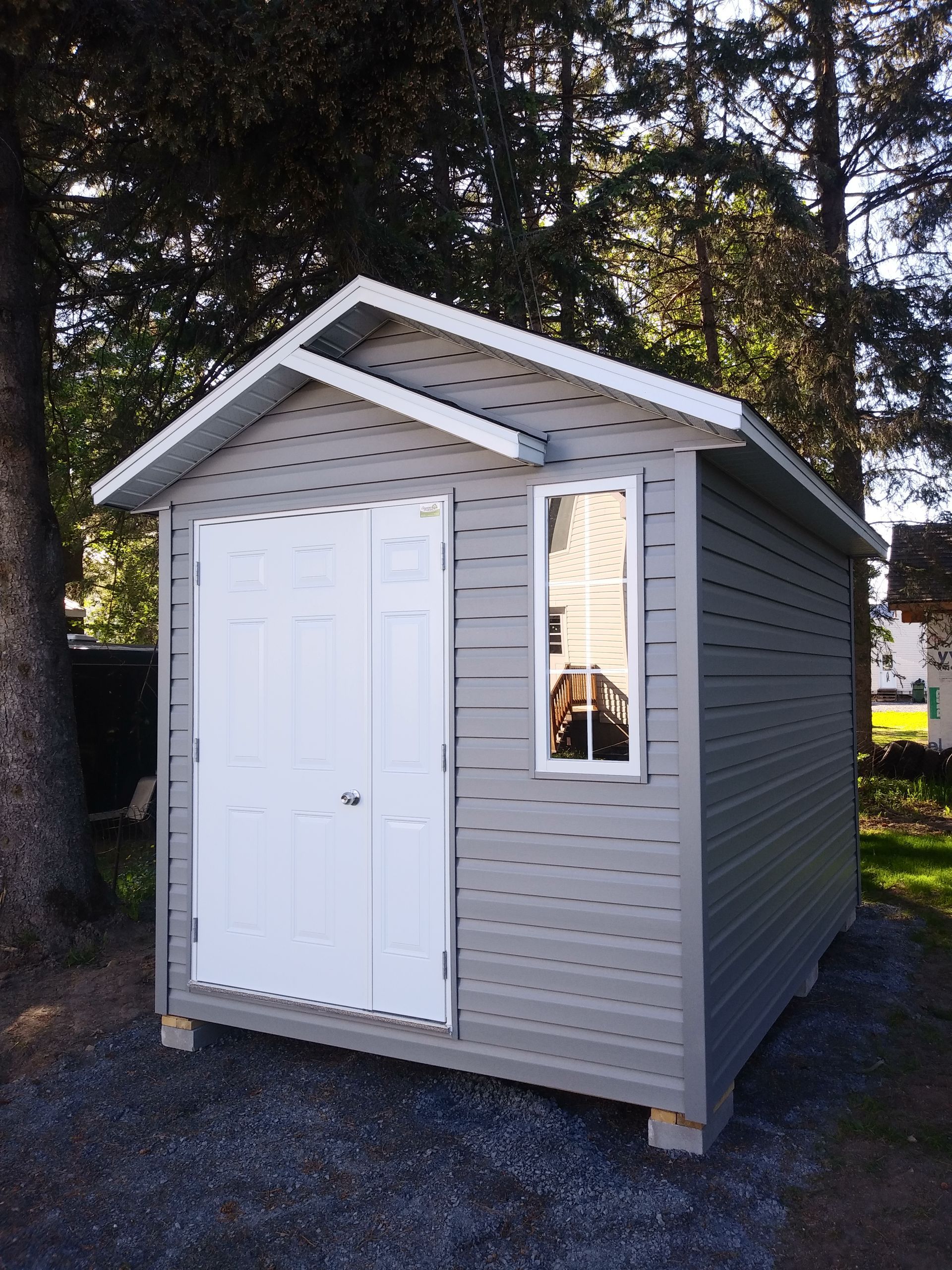 Abri de jardin gris avec portes et fenêtre blanches, sur parpaings dans une cour.
