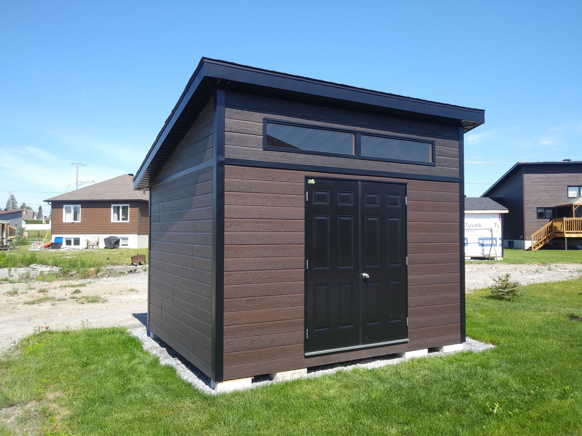 Abri de jardin marron et noir avec un toit en pente, des portes doubles et une fenêtre rectangulaire, par une journée ensoleillée.
