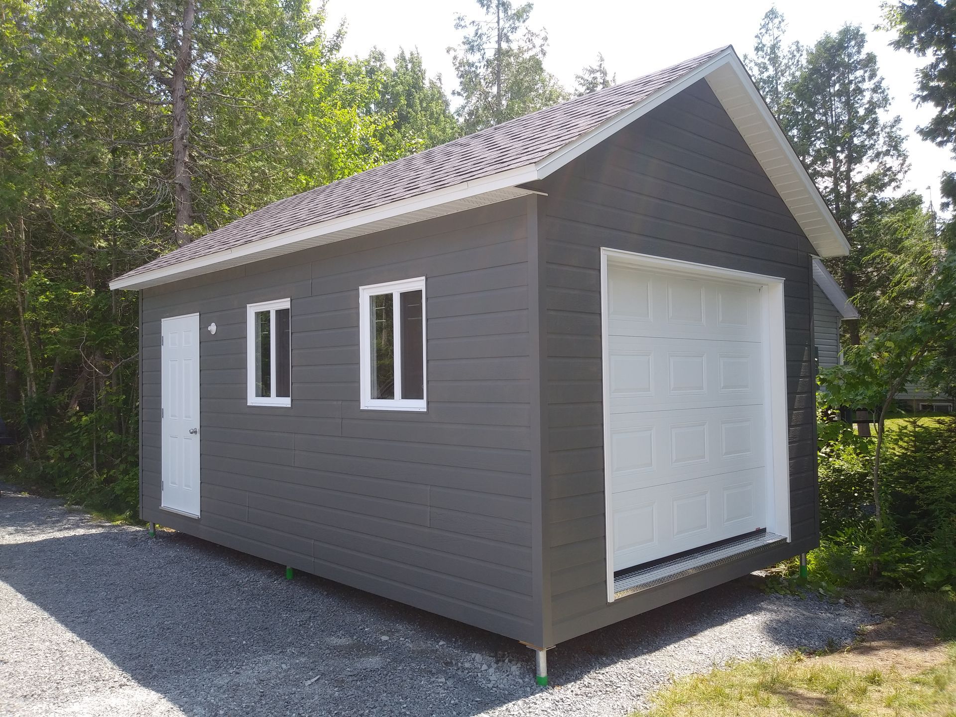 Abri de jardin gris avec porte, fenêtres et porte de garage blanches. Allée en gravier, arbres verts en arrière-plan.