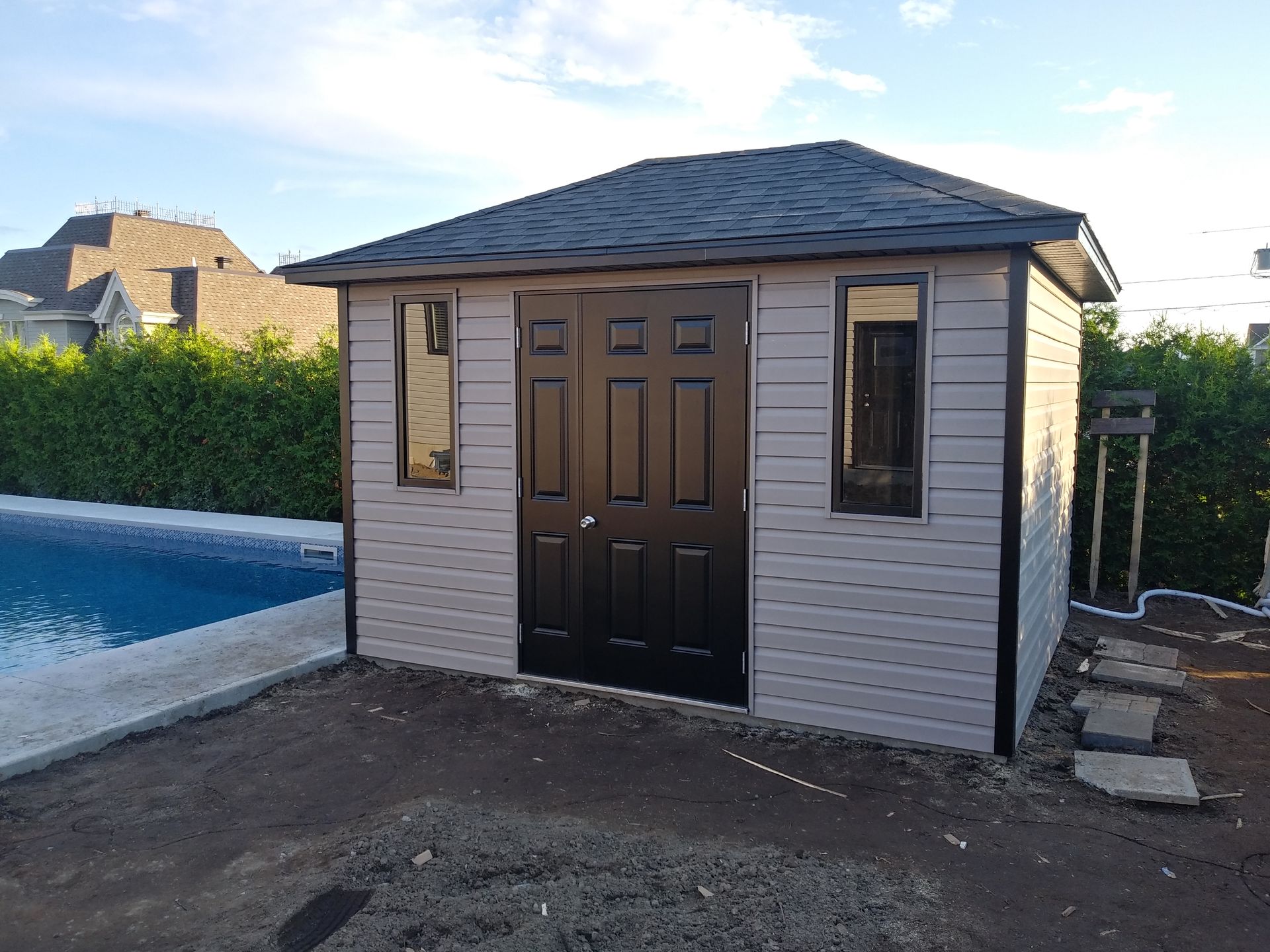 Abri de jardin beige avec portes et toit noirs, deux fenêtres, situé au bord d'une piscine par une journée ensoleillée.