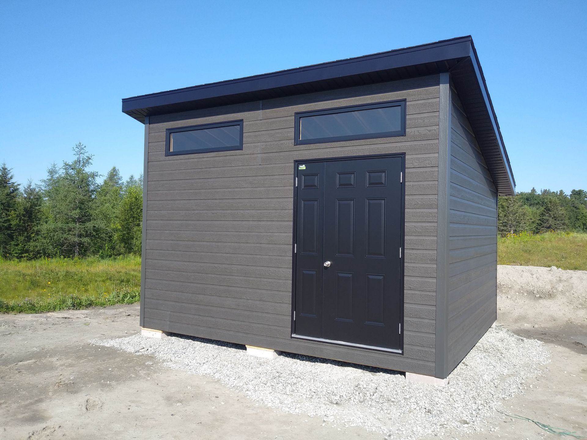 Cabane grise avec une porte noire et deux petites fenêtres sous un ciel bleu.