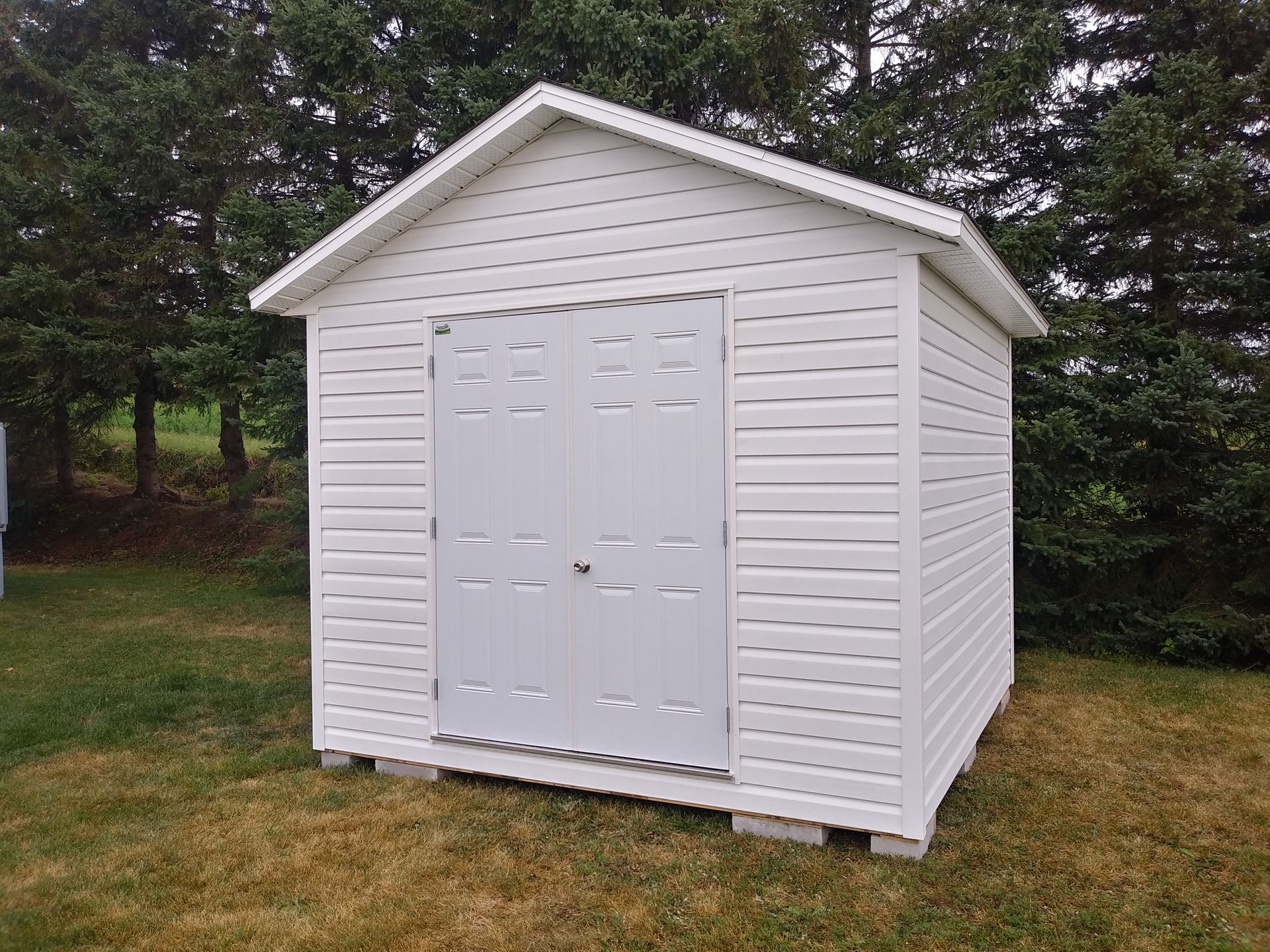 Abri de jardin blanc à double porte, situé dans une cour gazonnée et entourée d'arbres.